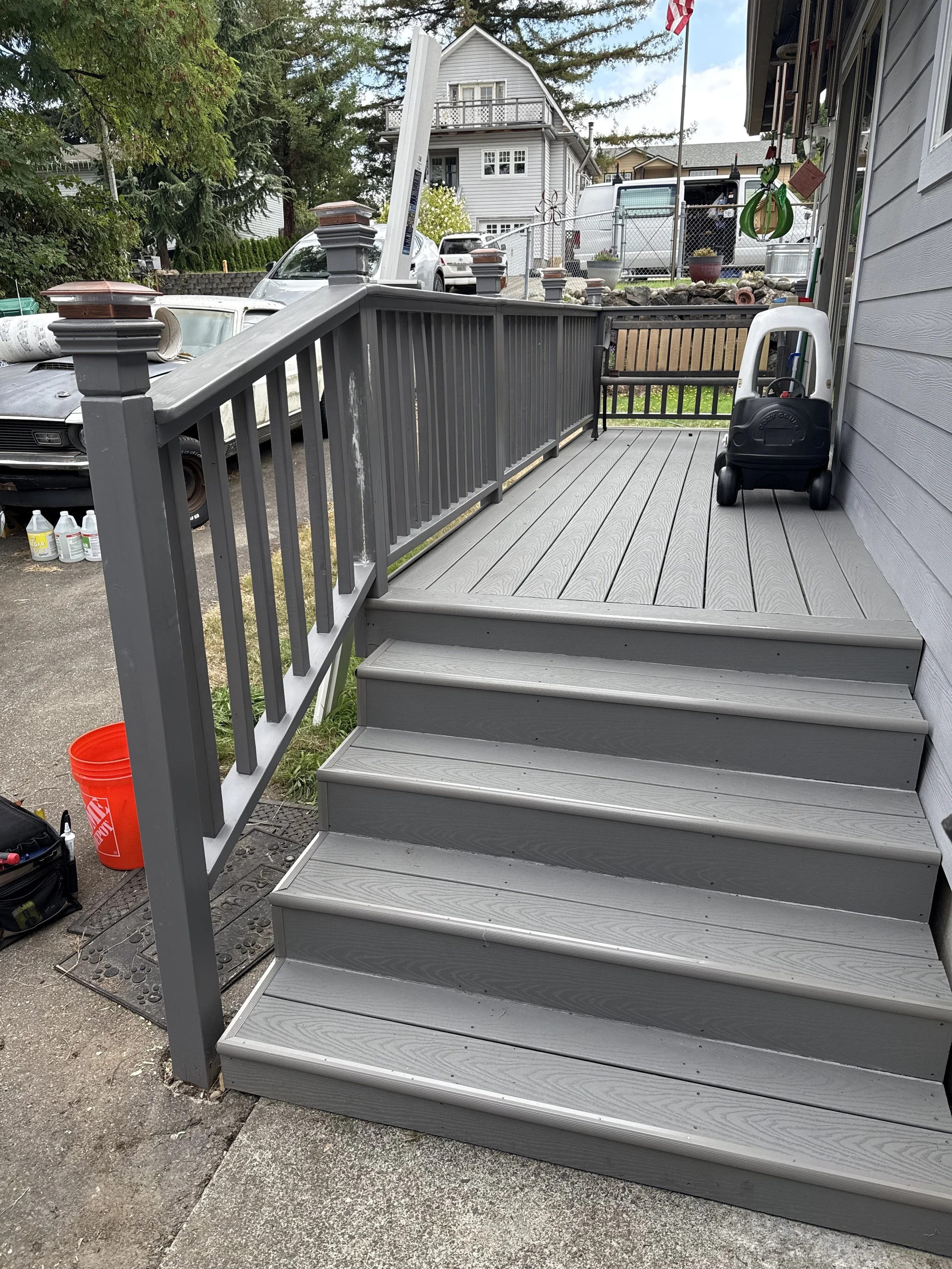 A concrete sidewalk and a small set of gray wooden stairs leading to a gray wooden deck with a gray railing. On the deck, there is a small black and white toy truck and some gardening tools hanging on the wall. The background shows a fence, a house,