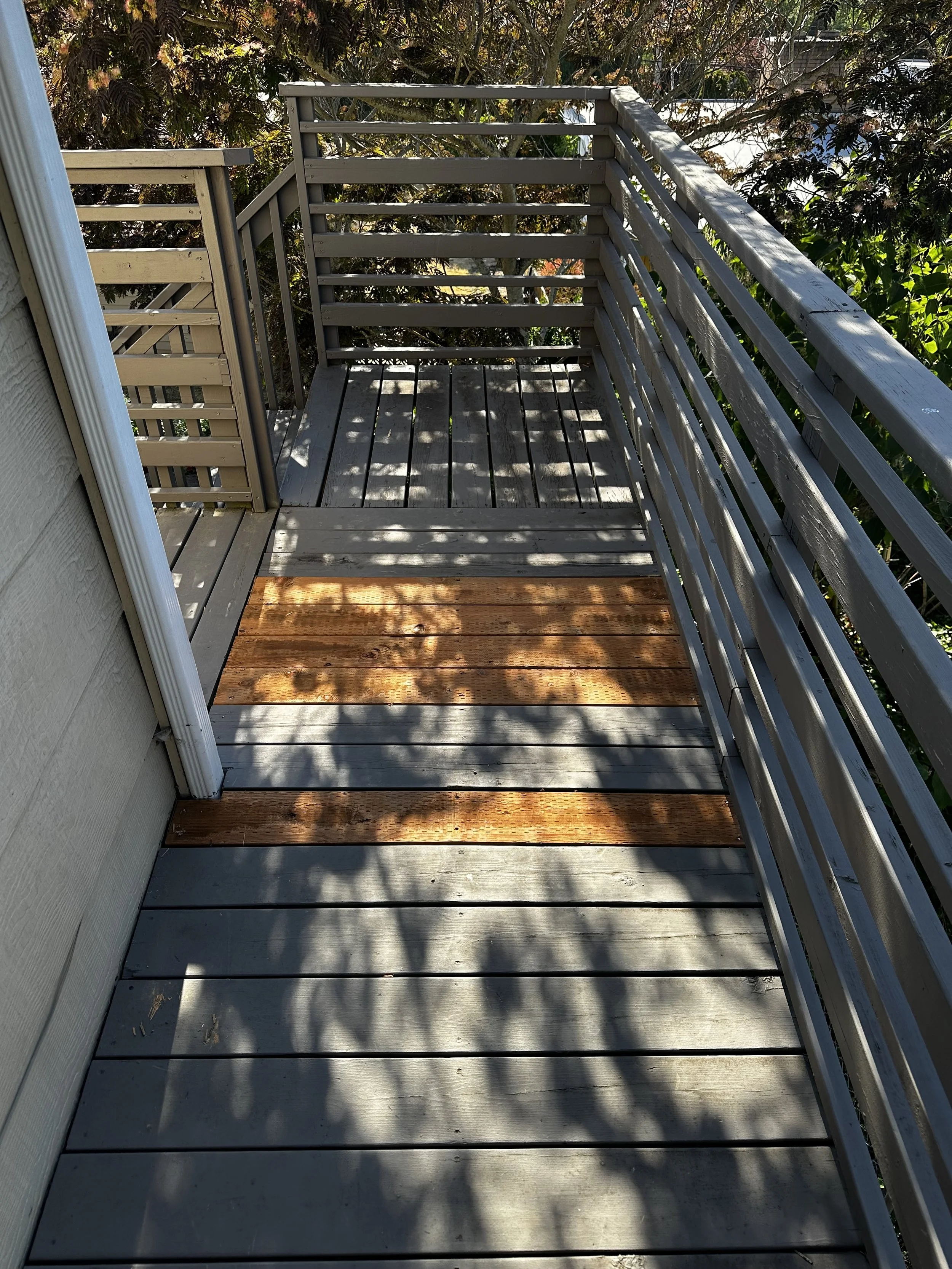 Small outdoor balcony with gray wooden floor and a few new wooden tiles, surrounded by a gray wooden railing, and shaded by nearby trees.