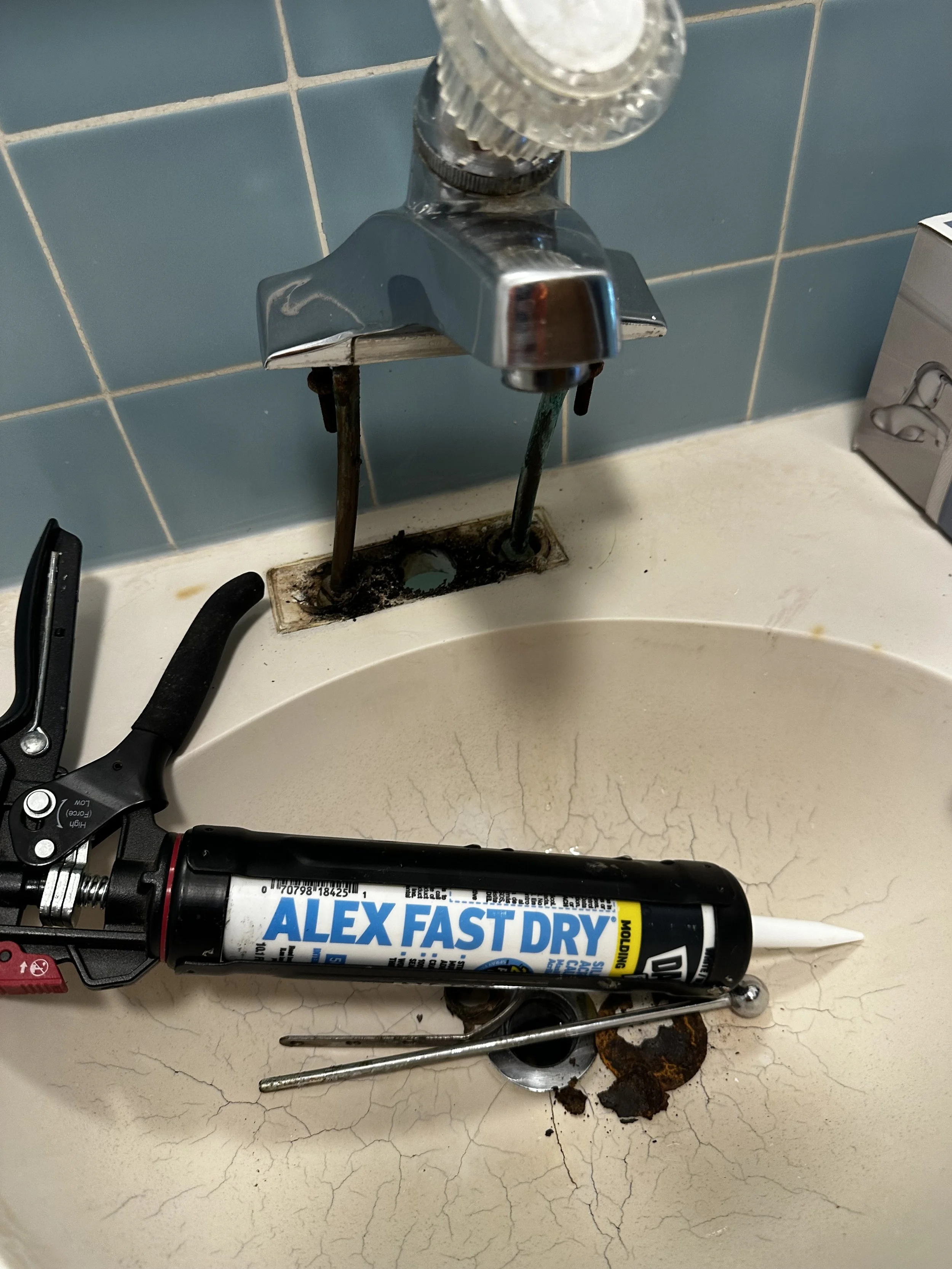 Bathroom sink with a partially removed faucet, a tube of Alex Fast Dry adhesive, a wrench, and some rust and debris around the drain.