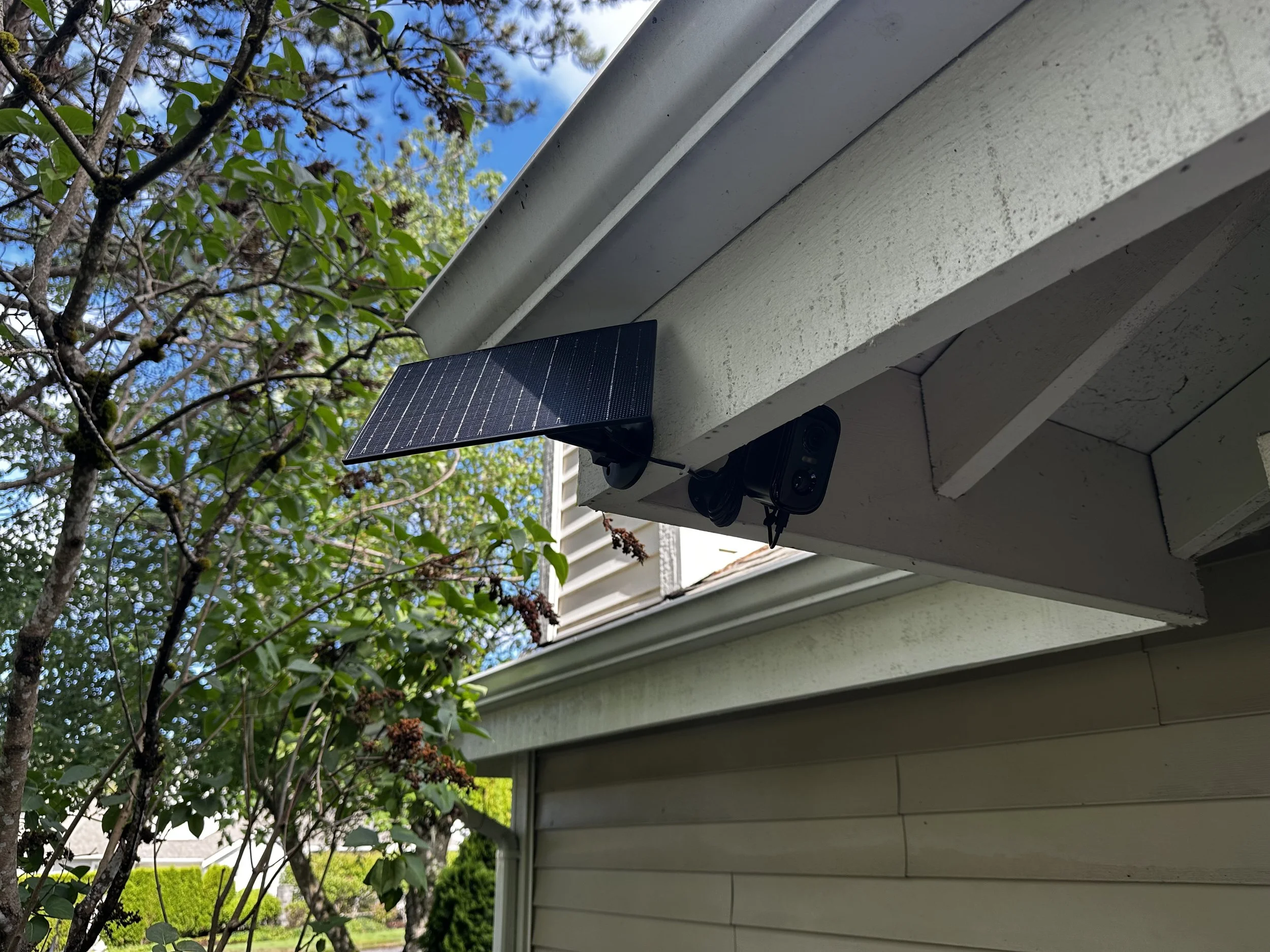 A small solar panel mounted on the edge of a house's roof near a security camera, with trees and blue sky in the background.