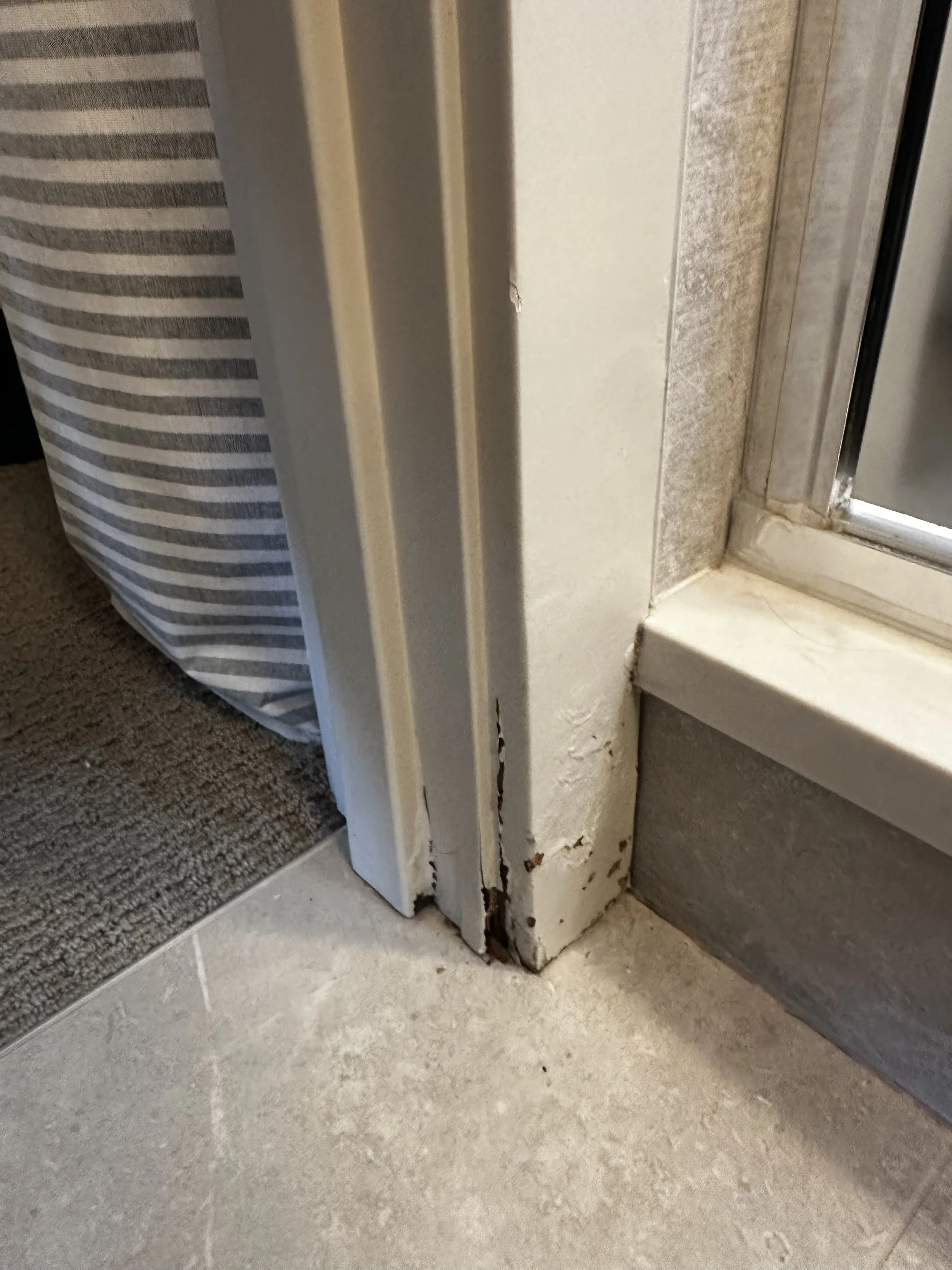Close-up of a door frame corner with peeling paint and damage at the bottom, next to a sliding glass door and patterned fabric in the background.