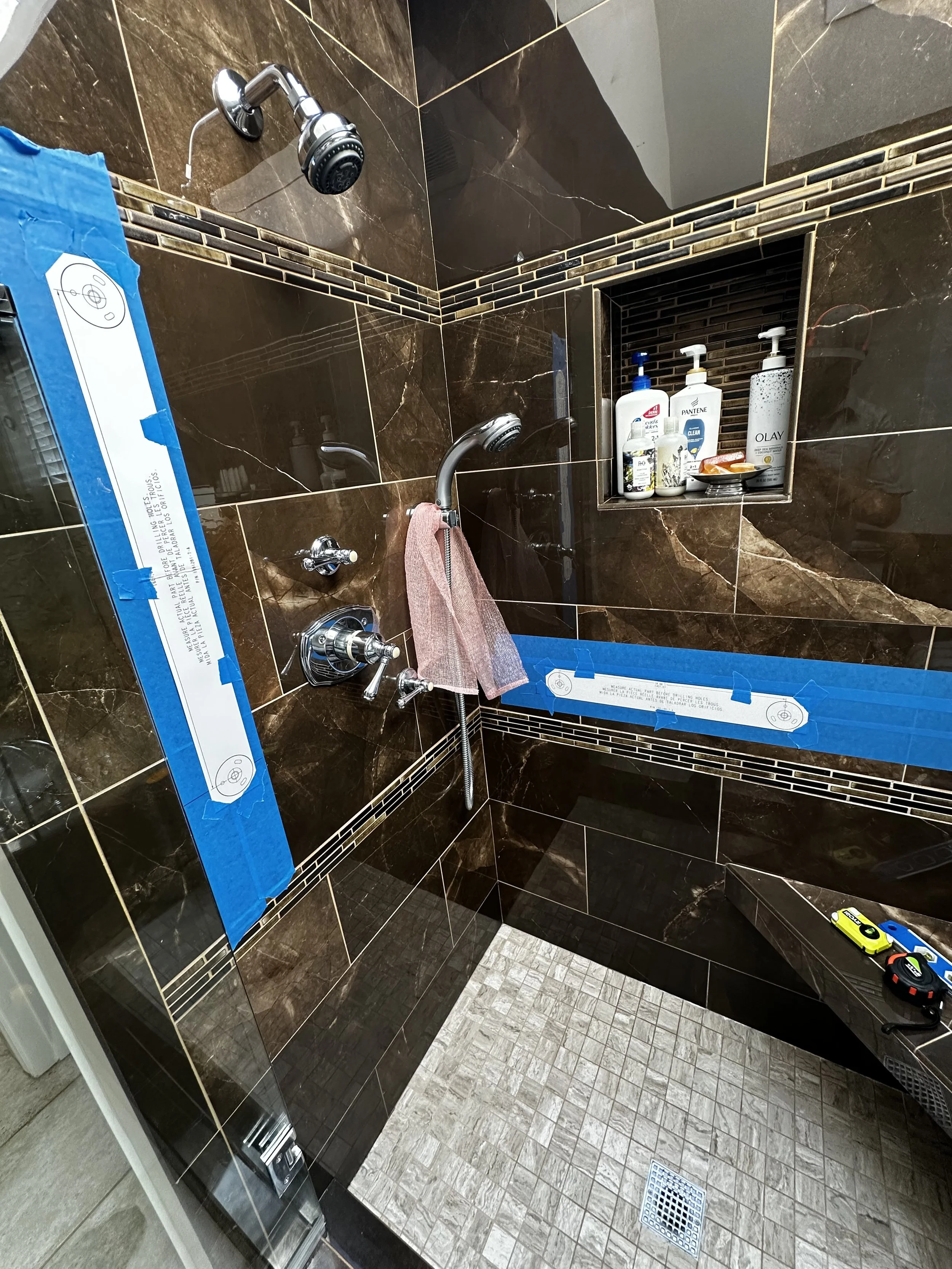 Brown tiled shower with two handheld shower heads, a built-in niche containing toiletries, a pink towel hanging on a metal bar, and blue painter's tape on the wall. There are some toys on the corner ledge inside the shower.