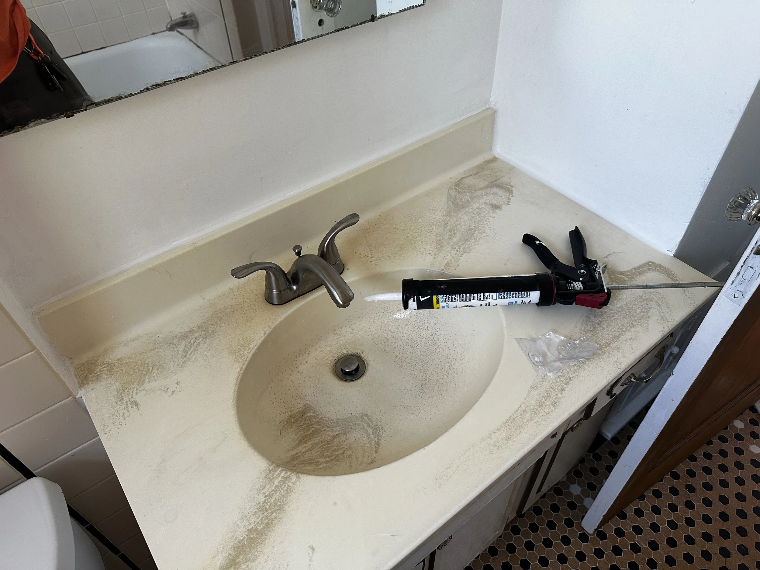 Bathroom sink with a dirty surface, a tube of sealant with a caulking gun placed on the countertop.