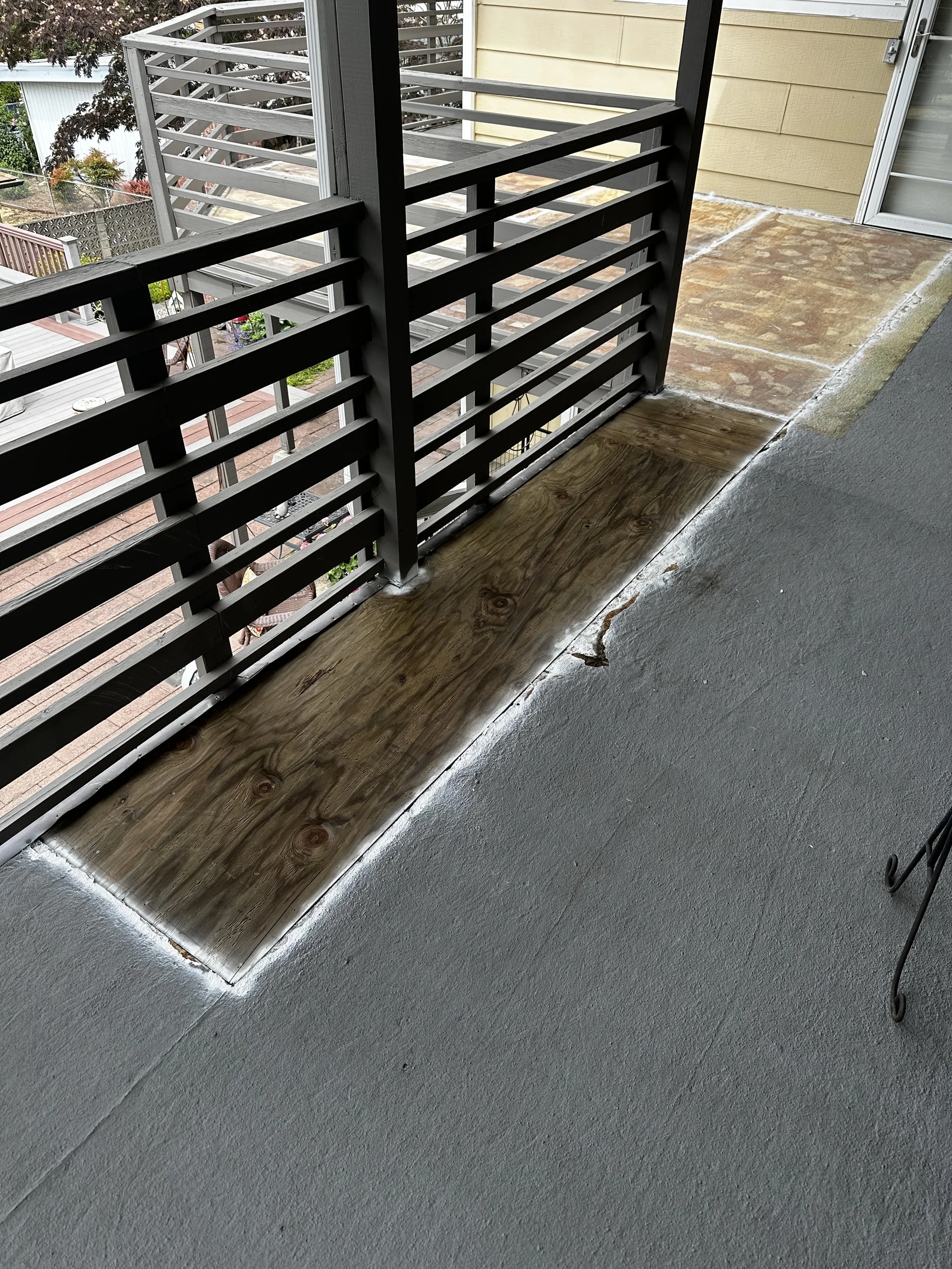 A balcony with a black metal railing, partially removed wooden flooring revealing the subfloor, and gray concrete flooring outside the door.