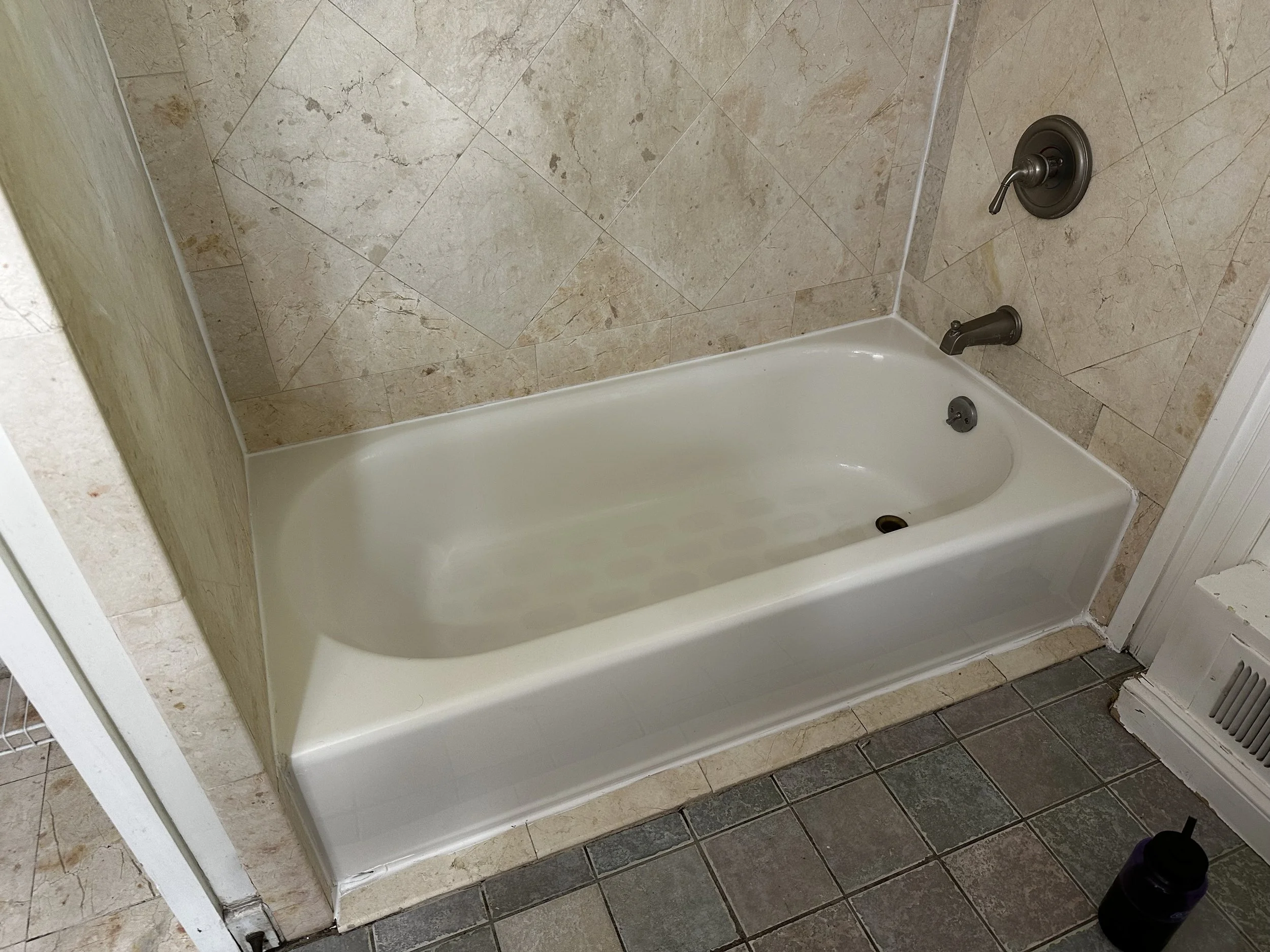 White bathtub with bronze fixtures, set against a beige tiled wall and floor, with a purple water bottle on the floor nearby.