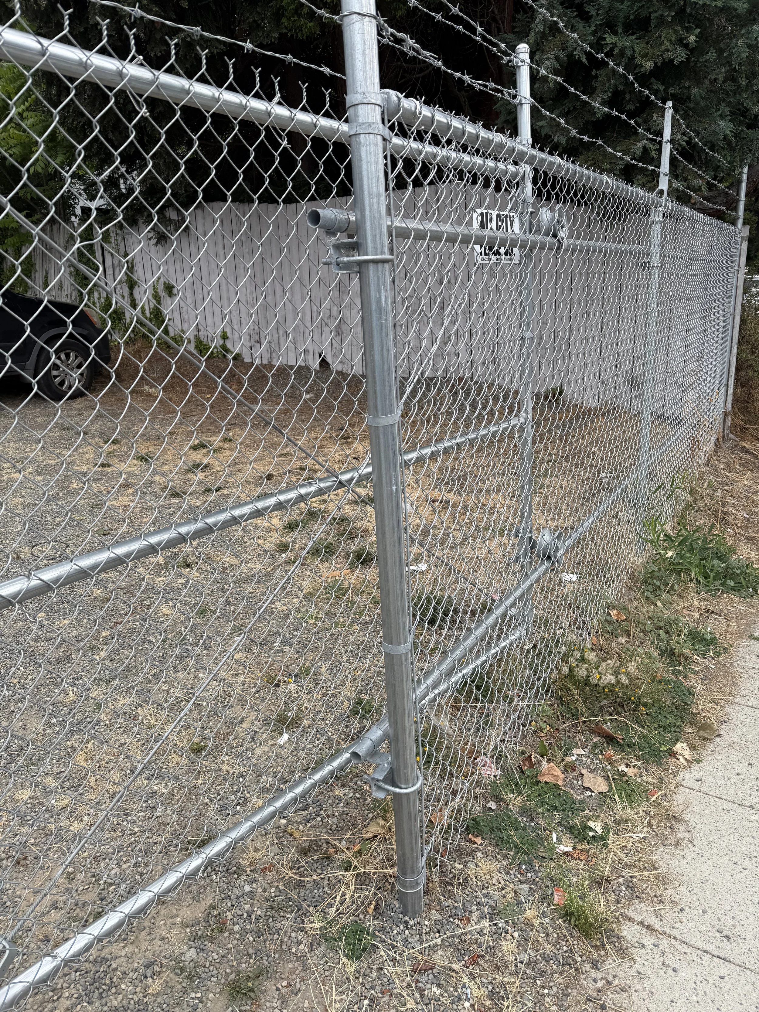 A chain-link fence with a gate and a sign that says 'City' and 'No Trespassing'. There is a car parked behind the fence and some trees in the background.