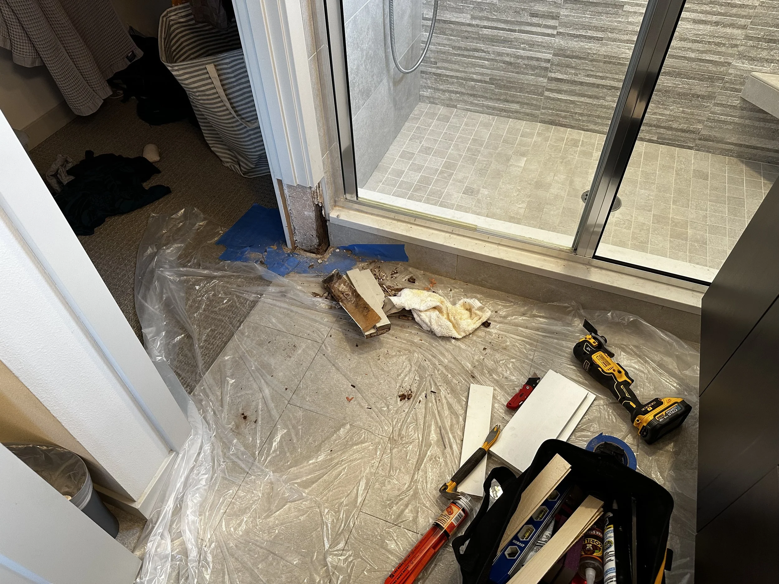 Construction or renovation scene showing a partially removed shower threshold or door frame, with tools and debris on the plastic sheet covering the floor, and a glass shower door leading to a tiled shower area.