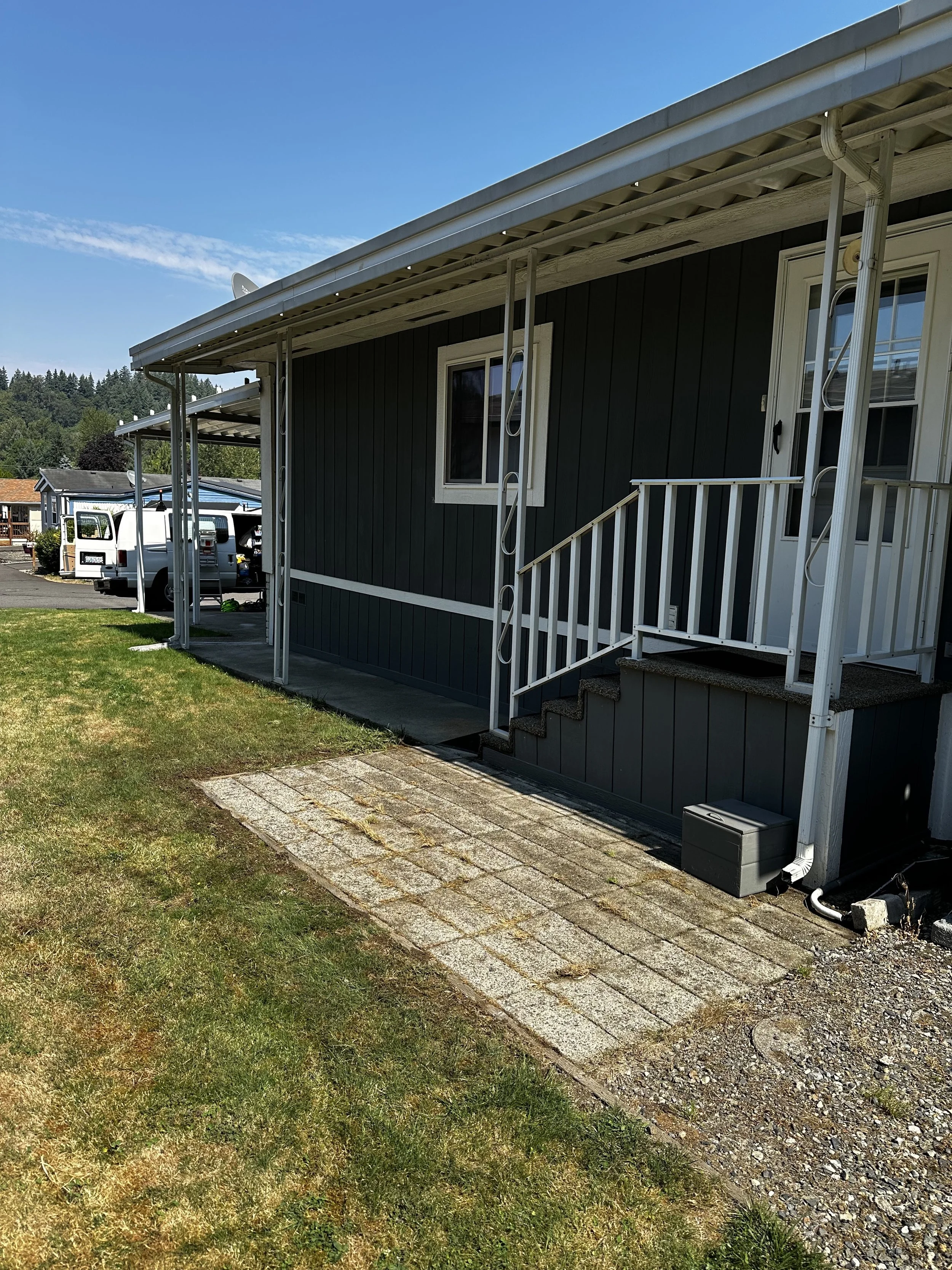 Front view of a house with steps leading to the door, a small porch, and a paved walkway. There are white gutters and siding, with a grassy yard in the foreground and a clear blue sky.