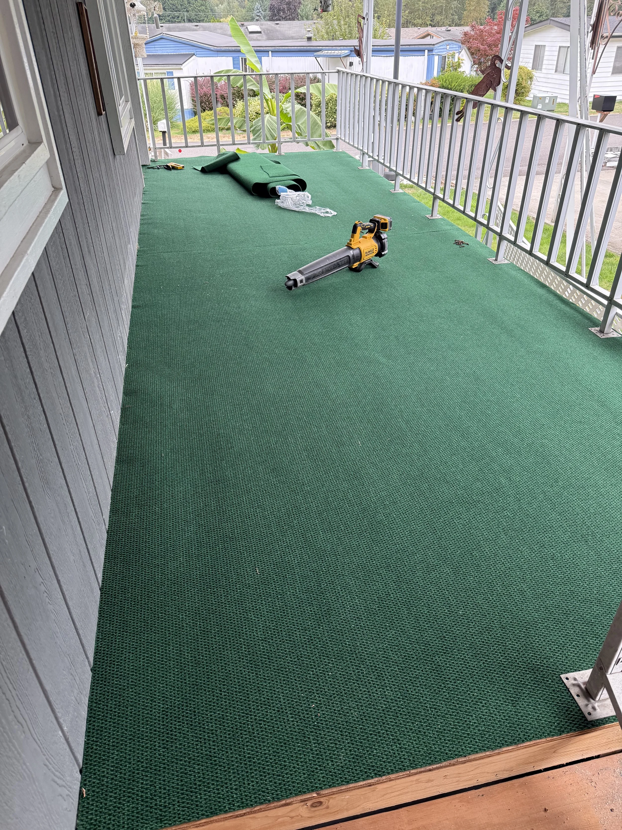 A balcony with green outdoor carpet, tools, and a leaf blower, overlooking a neighborhood with houses and trees.