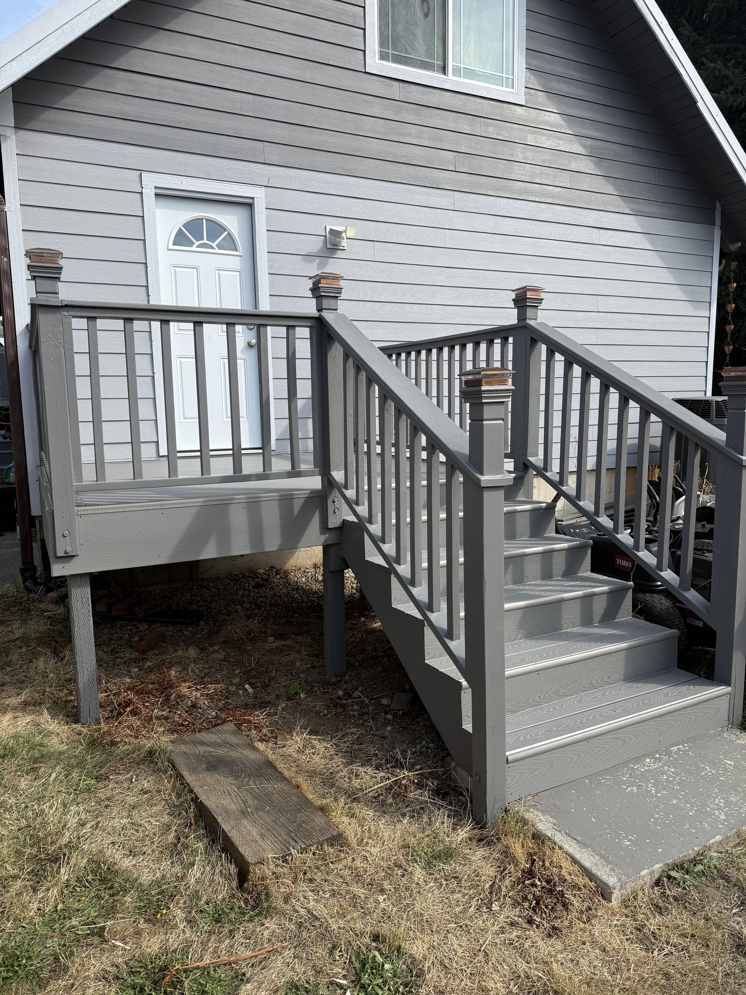 Gray wooden staircase leading to a back door of a house with gray siding and a small landing. The stairs have railings on both sides and decorative posts with tan caps. There is a small concrete pad at the base of the stairs and a patch of grass with