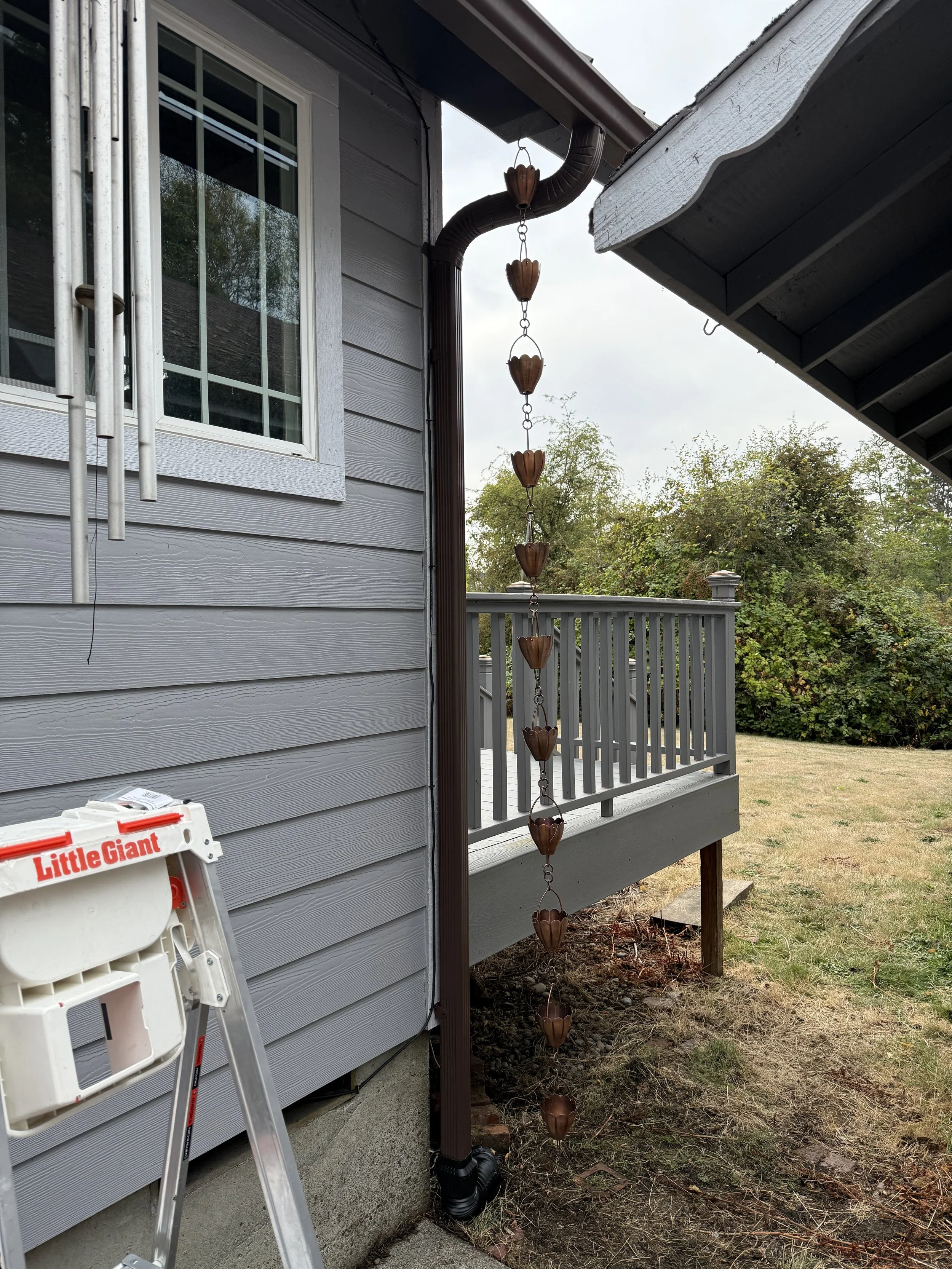 A gray house with a window, a gutter system, and a hanging decorative rain chain made of butterfly-shaped copper cups. A ladder marked 'Little Giant' is in the foreground and a wooden deck with a railing is in the background.