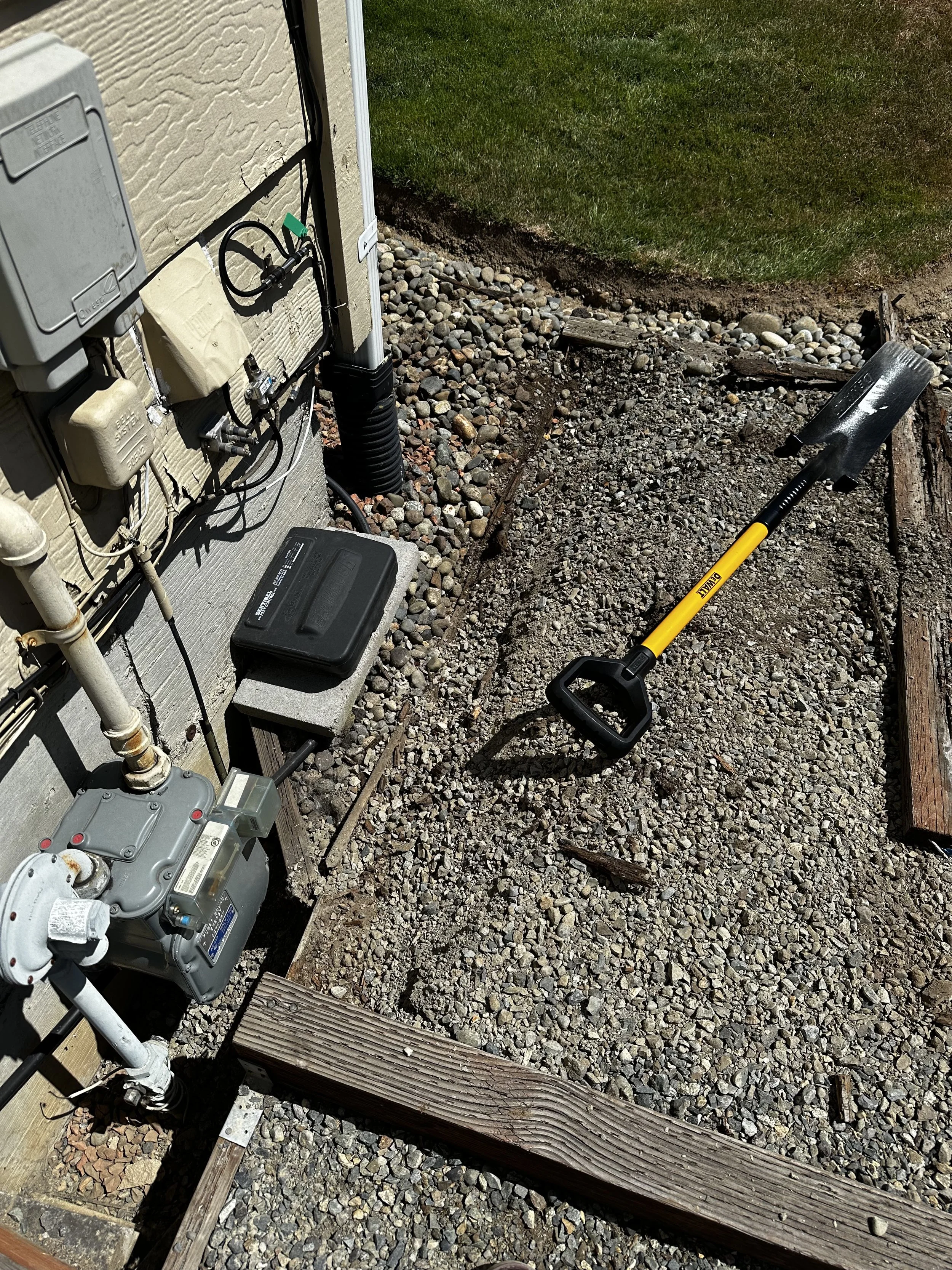 A gardening shovel with a yellow handle and black tip lying on a gravel-covered ground outside a house. There is a small wooden border along one side, and exterior electrical wiring and equipment attached to the house wall.