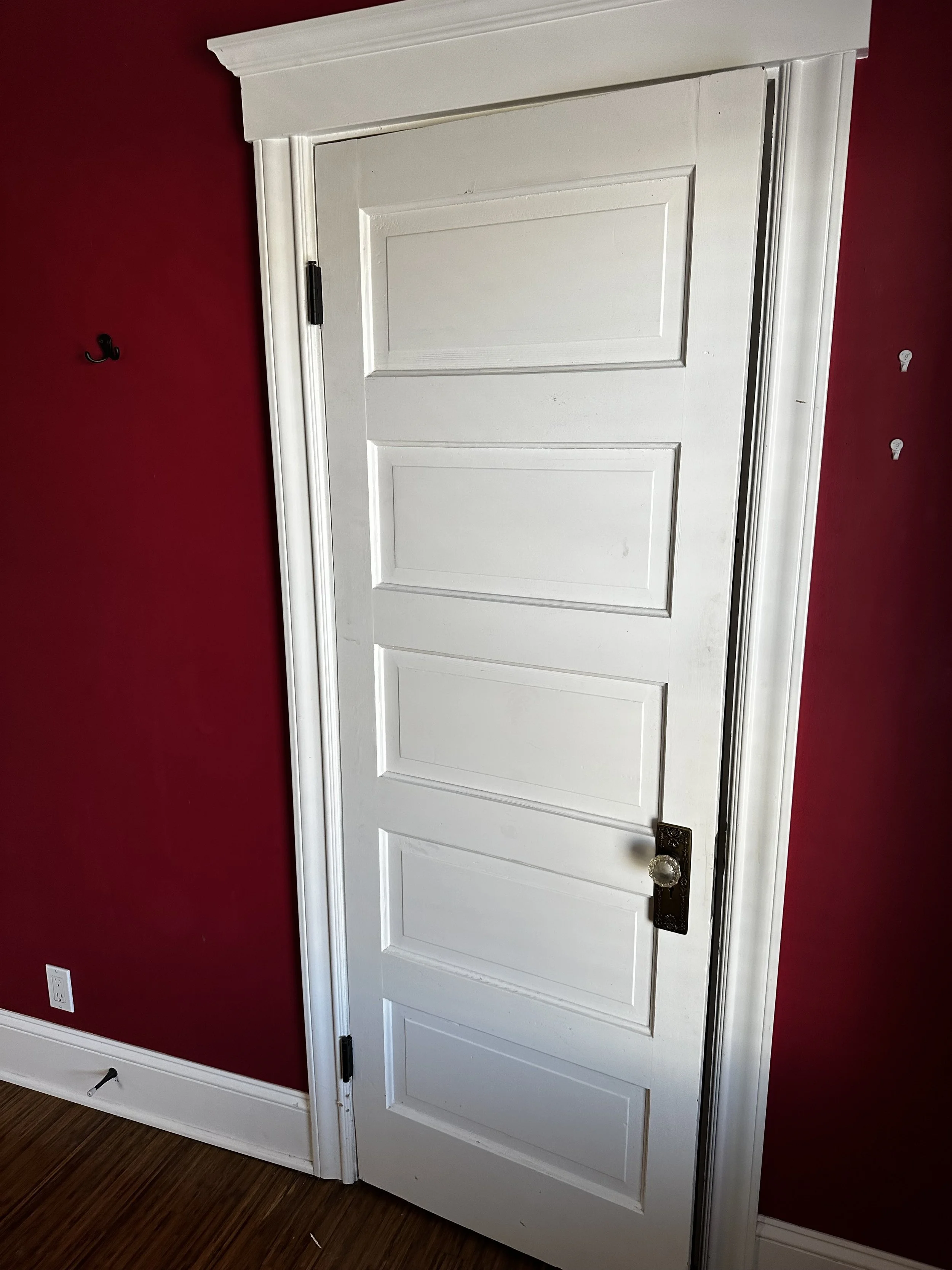 White interior door with four raised panels and a vintage brass doorknob, set in a room with red walls and dark wood flooring.