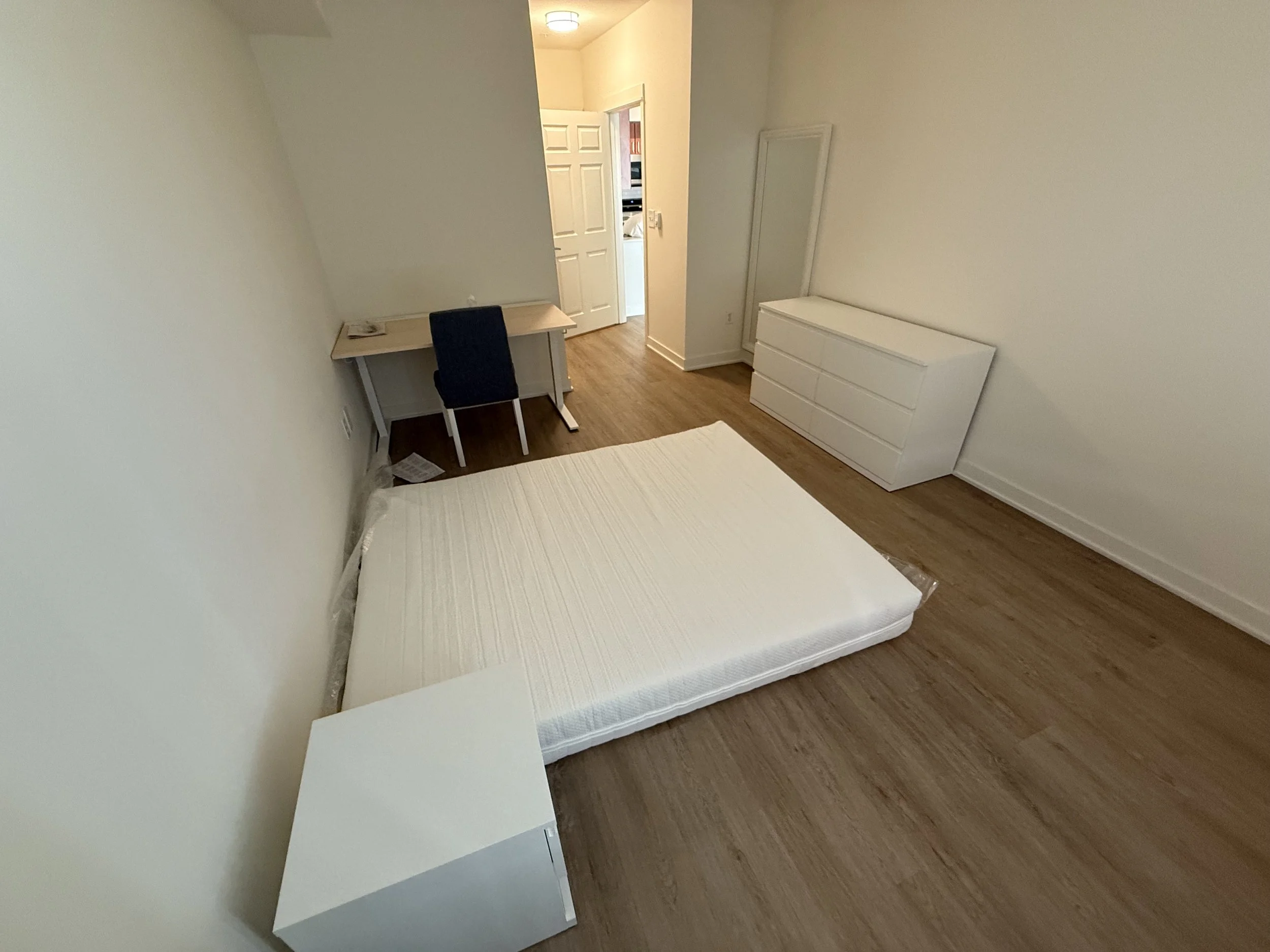 Empty bedroom with a mattress on the floor, a white nightstand, a desk with a black chair, and a white dresser against the wall. The walls are white and the floor has wood flooring.