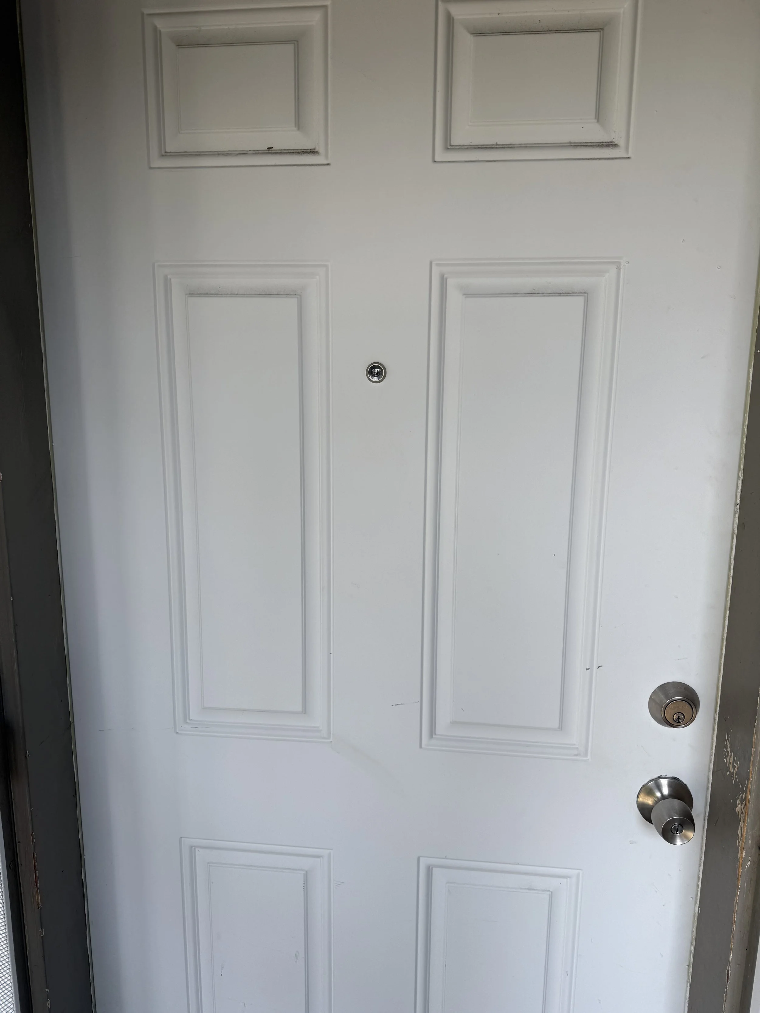 Close-up of a plain white exterior door with a peephole, a door handle, and a deadbolt lock, surrounded by a dark-colored door frame.