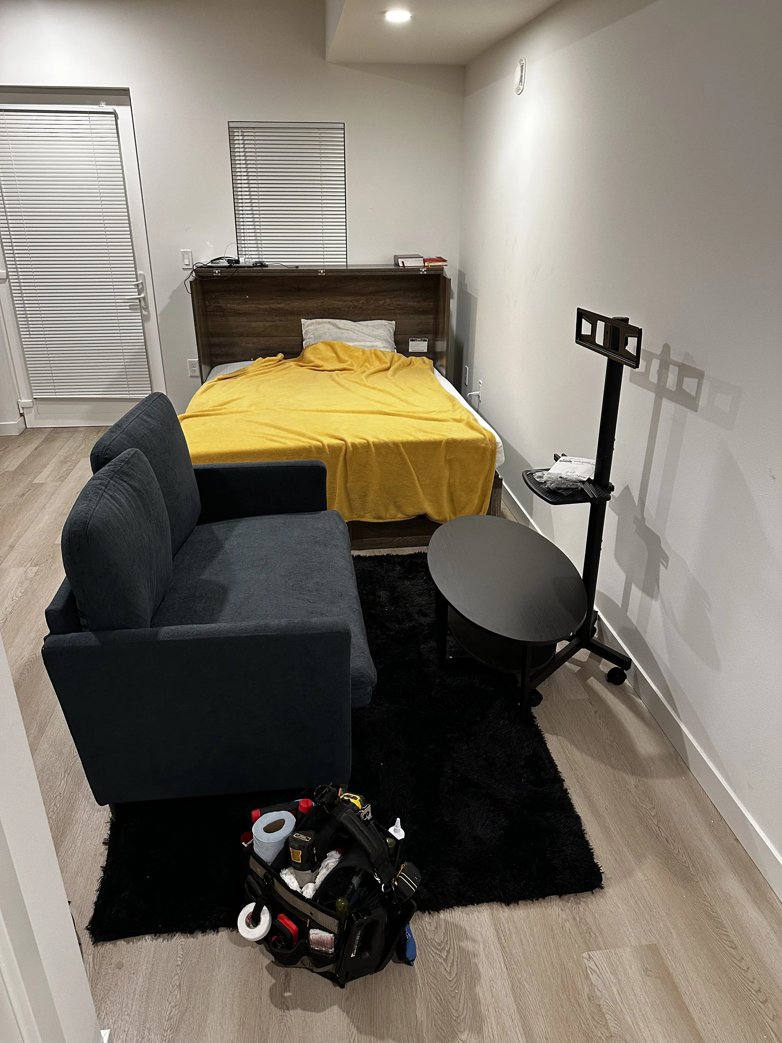 A small living space with a bed covered in a yellow blanket, a dark gray sofa, a small black coffee table, and a white wall-mounted TV bracket. There is a black tool bag with tools on the floor and a black area rug on light wood flooring. A large win