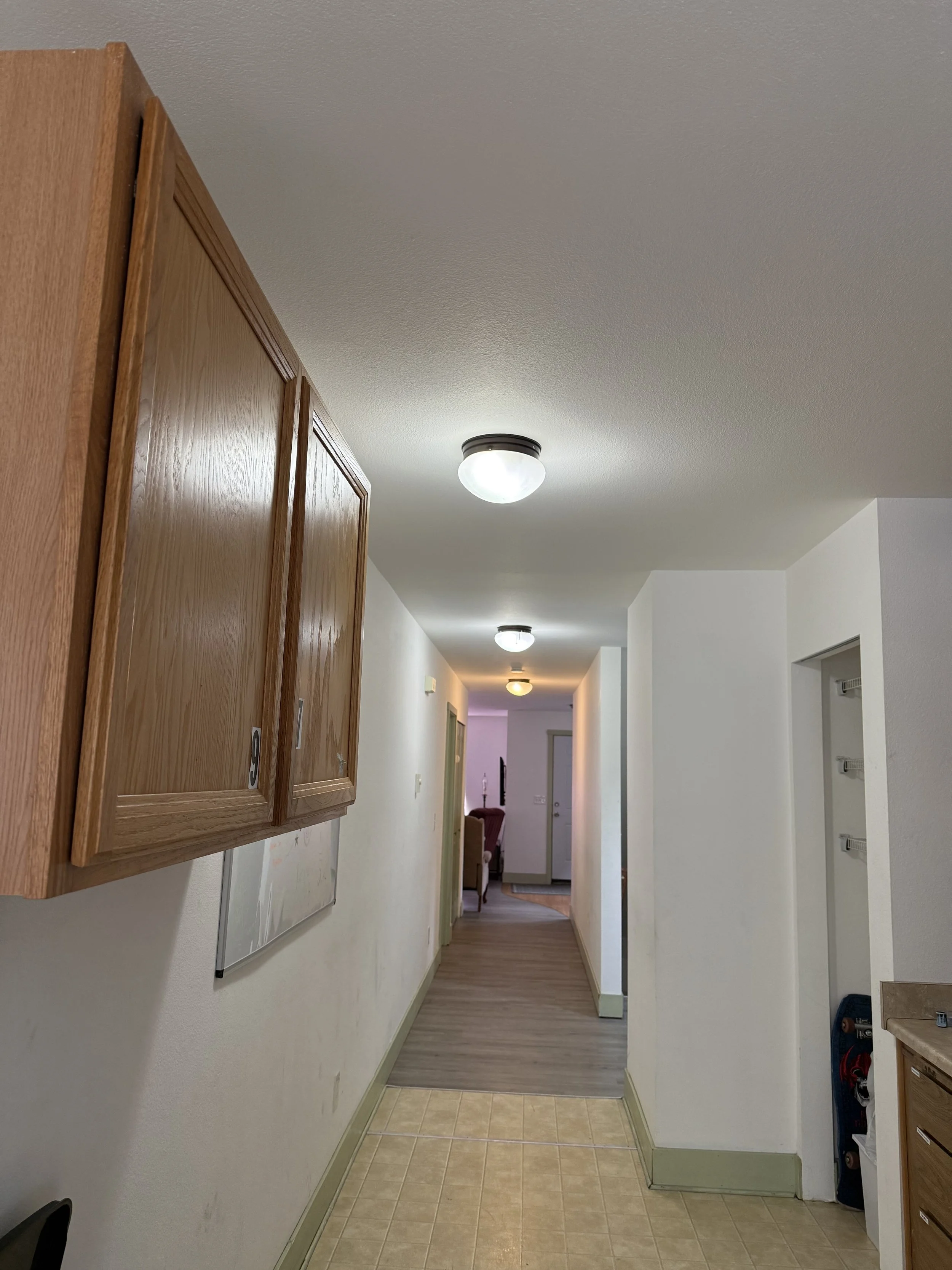 A hallway with wooden cabinetry on the left, white walls, a tiled floor near the camera, and a wood floor further down the hall. There are ceiling lights and a glimpse of a living area with furniture at the end of the hallway.