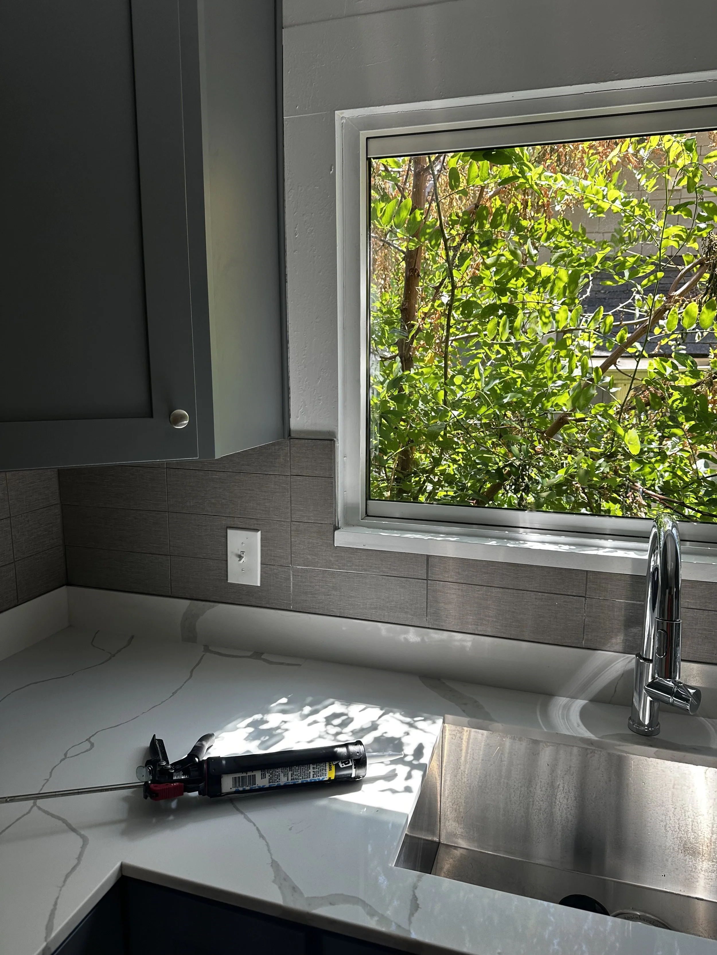 Kitchen countertop with caulking gun placed next to a sink, window overlooking green foliage outside.