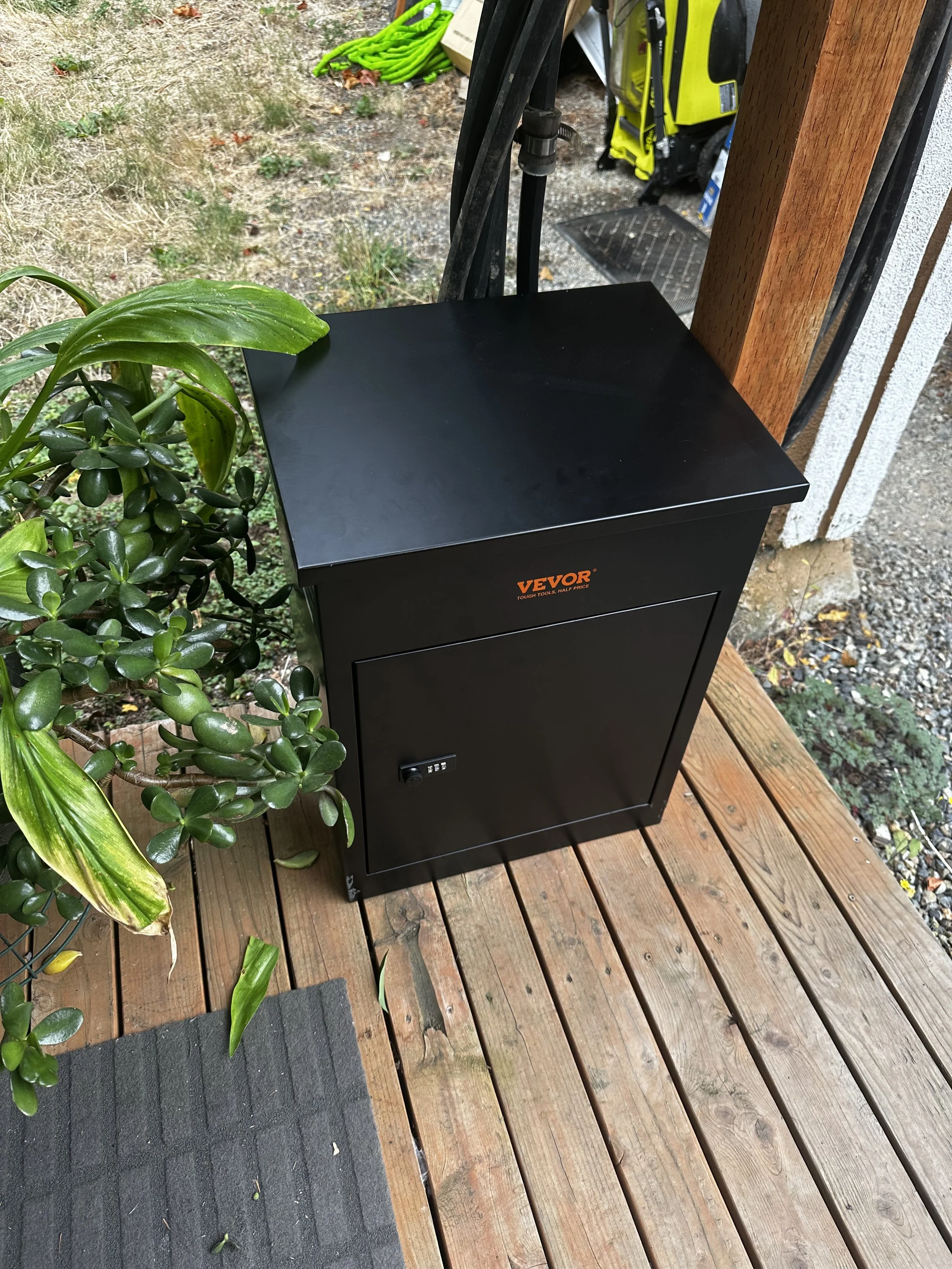 A black Secure Storage cabinet with a lock, placed on a wooden porch next to a green plant, outside a house.