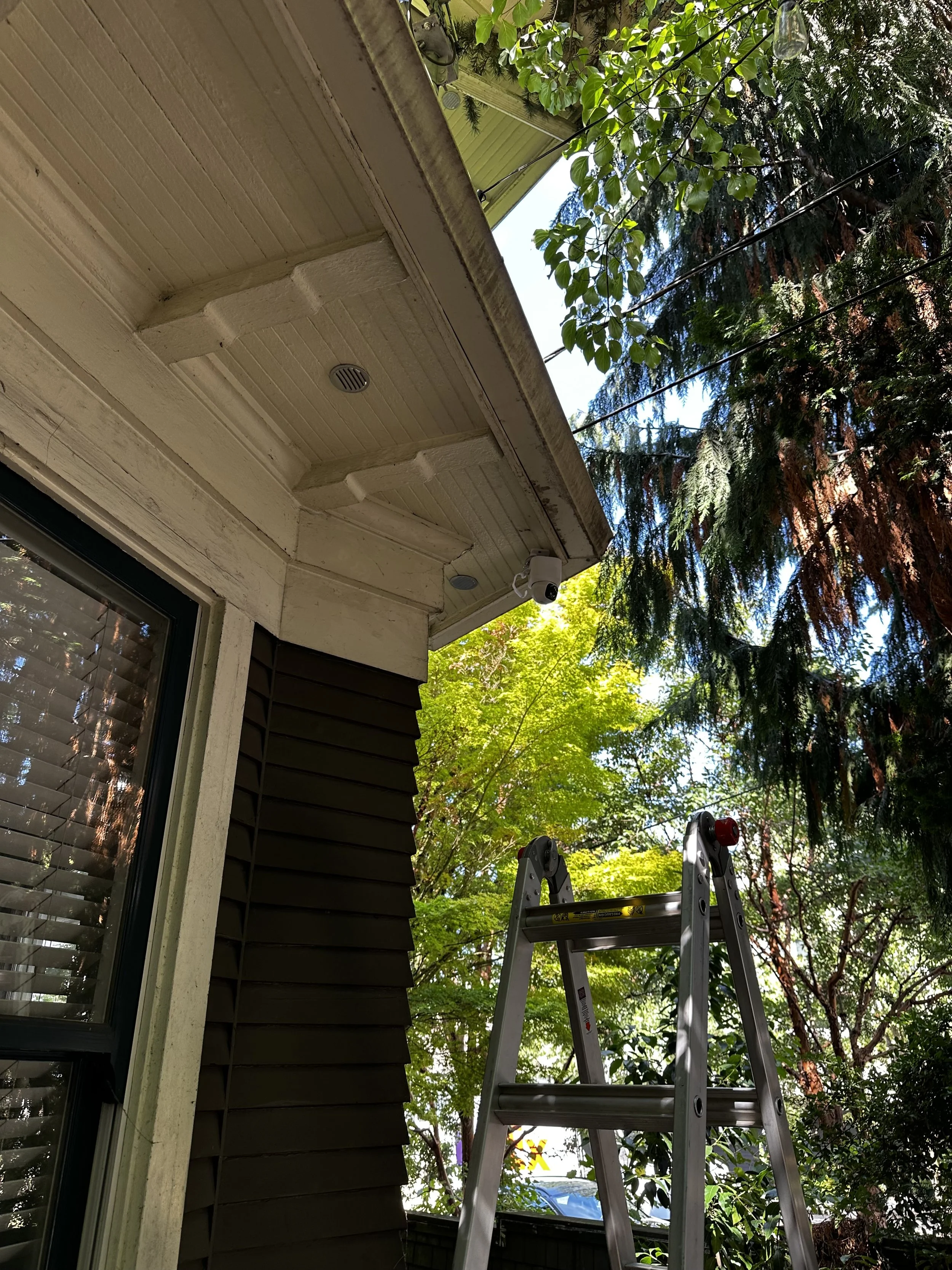 Exterior of a house with a ladder, trees, and a security camera under the eaves of the roof.