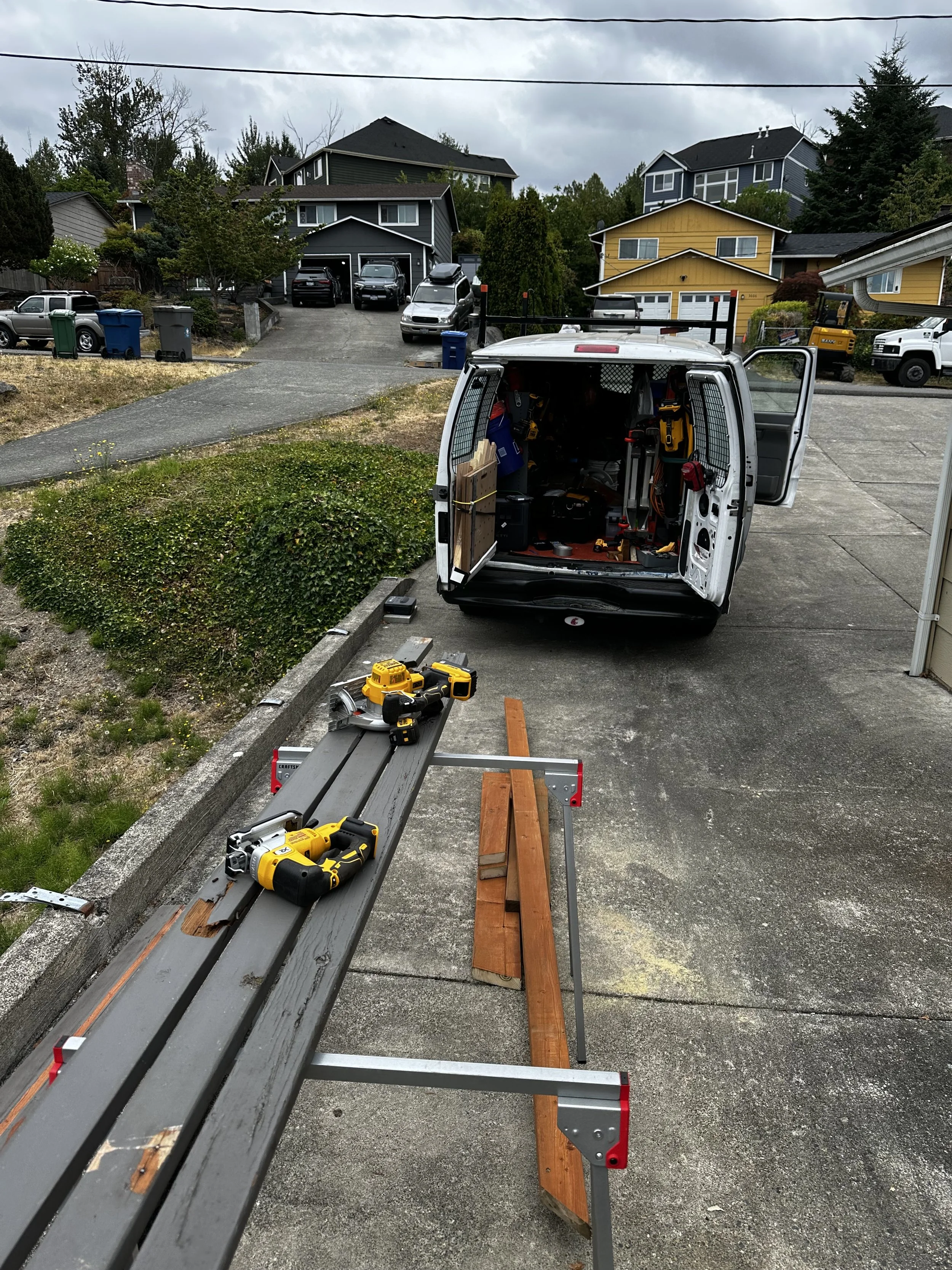 A work van with its rear doors open, parked in a driveway. Inside the van are various tools and equipment. A workbench with power tools is set up outside the van. The background shows a neighborhood with houses, trees, and parked cars.