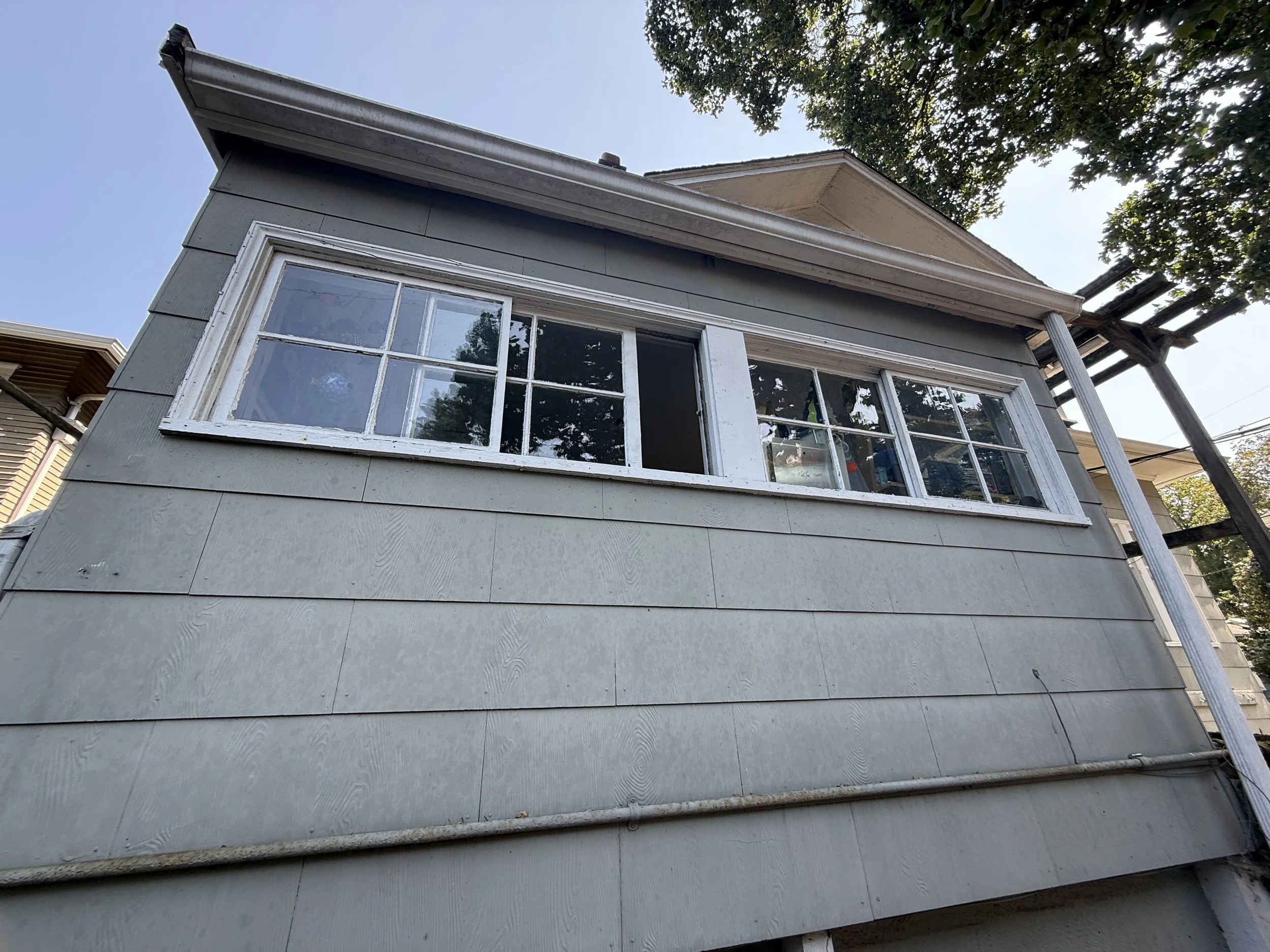 The photo shows the side of a house with light blue siding and a large multi-pane window. The house has a sloped roof and a small section of the neighboring house is visible. There is a tree with leaves and branches in the upper right corner, and a p