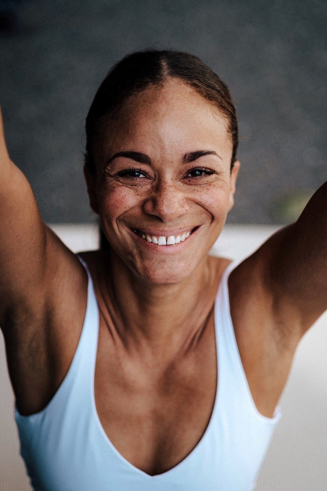 Lächelnde Frau mit braunen Haaren in einem weißen Top, die Arme hochhebt