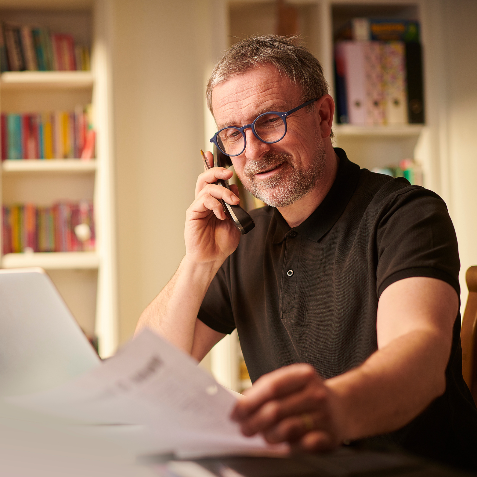 A middle-aged man with glasses and a beard, wearing a black polo shirt, is sitting at a desk talking on a landline phone and looking at some papers.