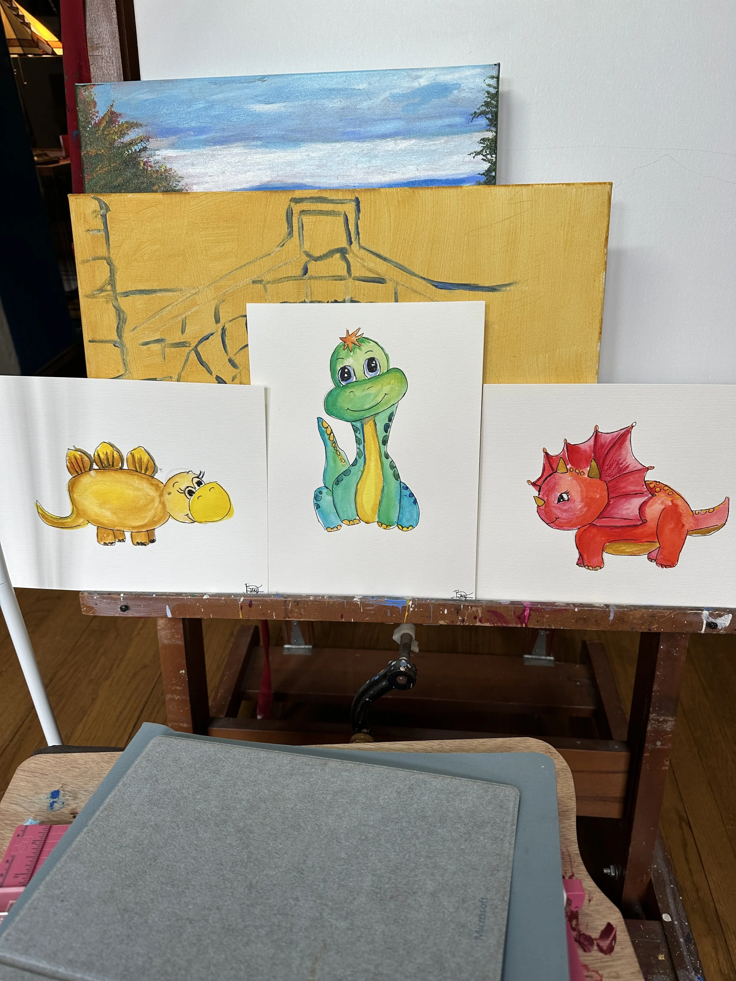 A display of colorful cartoon dinosaur paintings on white canvases, with a partially painted landscape in the background. The artworks feature a yellow dinosaur with orange tips, a green and blue dinosaur with a star on its head, and a red dinosaur with pink wings.