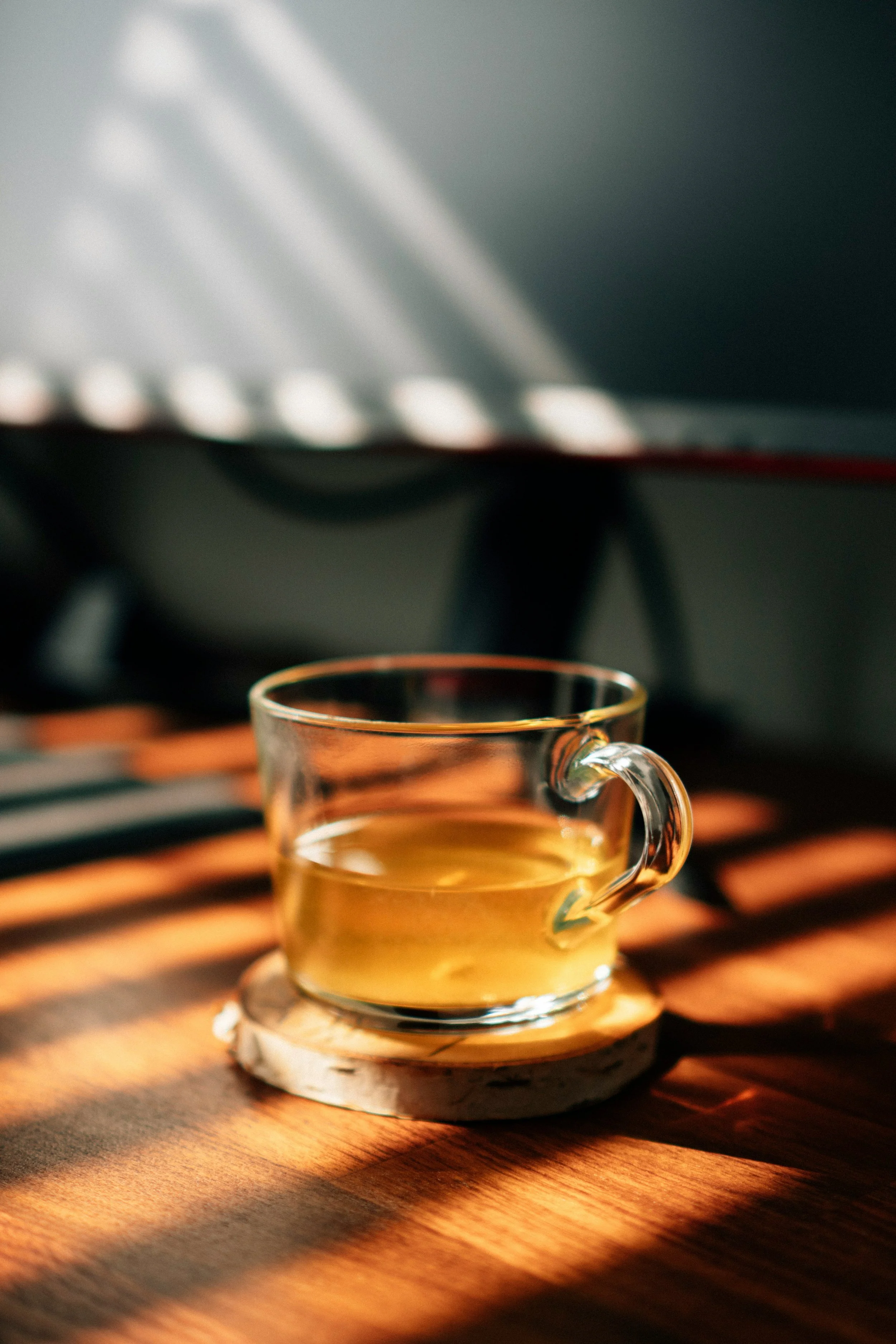 A clear glass cup with a small handle filled with a yellowish liquid sitting on a round coaster on a wooden surface, with light and shadow patterns across the scene.