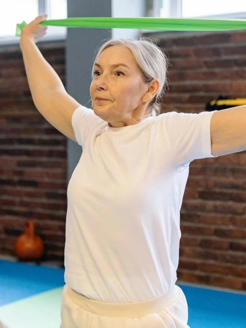 Older woman stretching with a green resistance band in a gym or fitness studio.