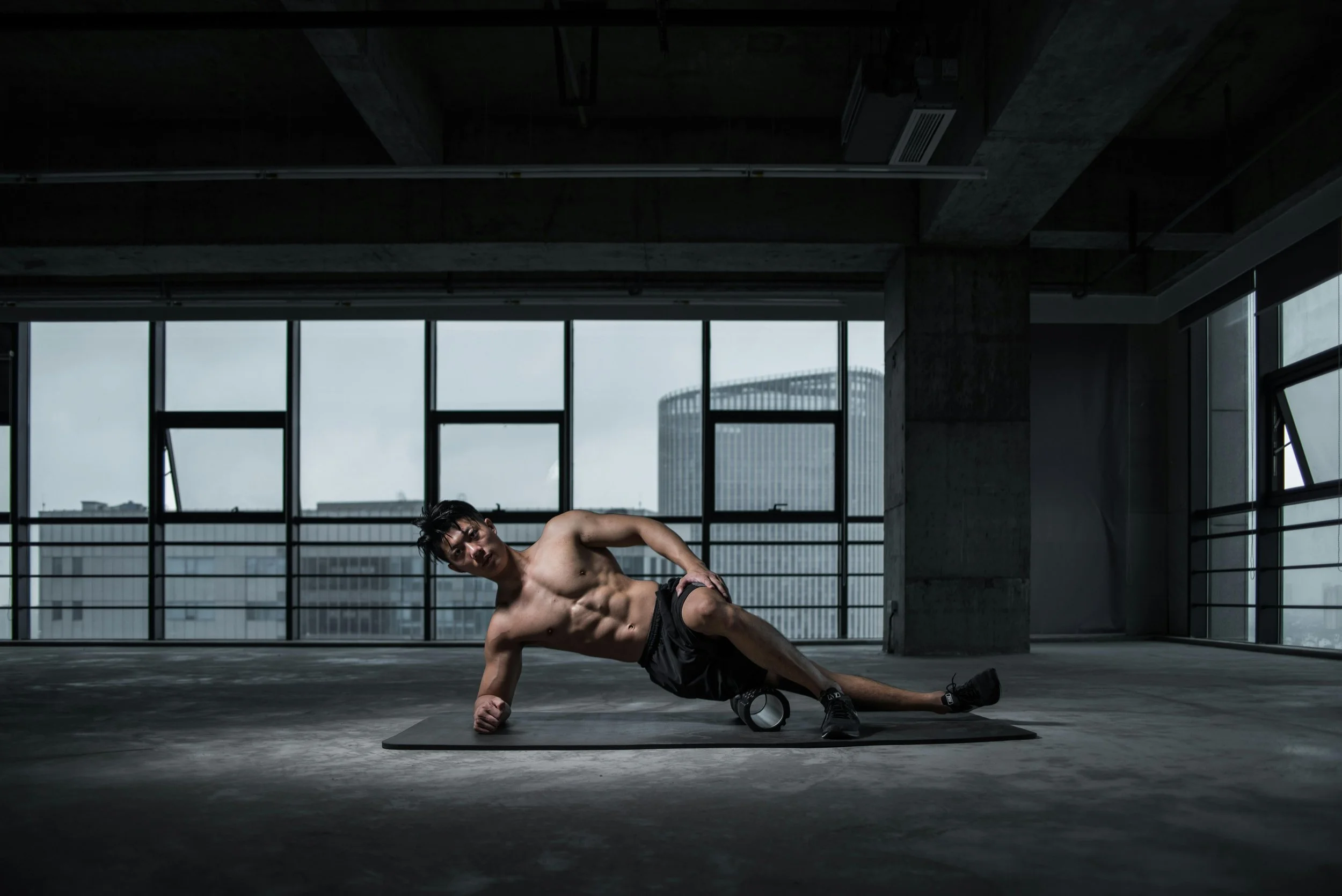 A shirtless man with a muscular physique doing a fitness exercise on a mat in an industrial-style gym with large windows and cityscape view.