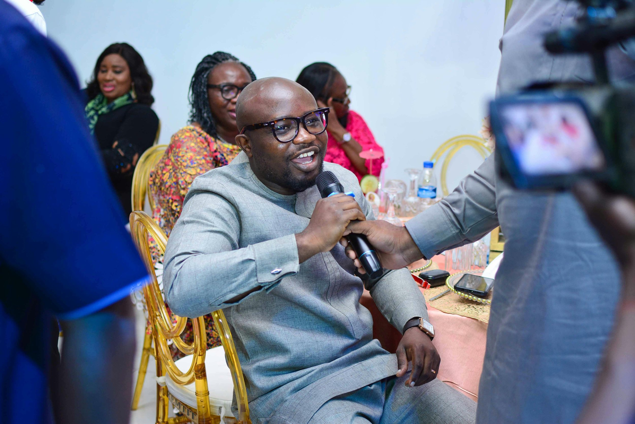 A smiling man in glasses speaks into a microphone during an event, while others seated around him look on attentively. Reflecting the perspective of Matt Arlyn on strategies for policy resilience.