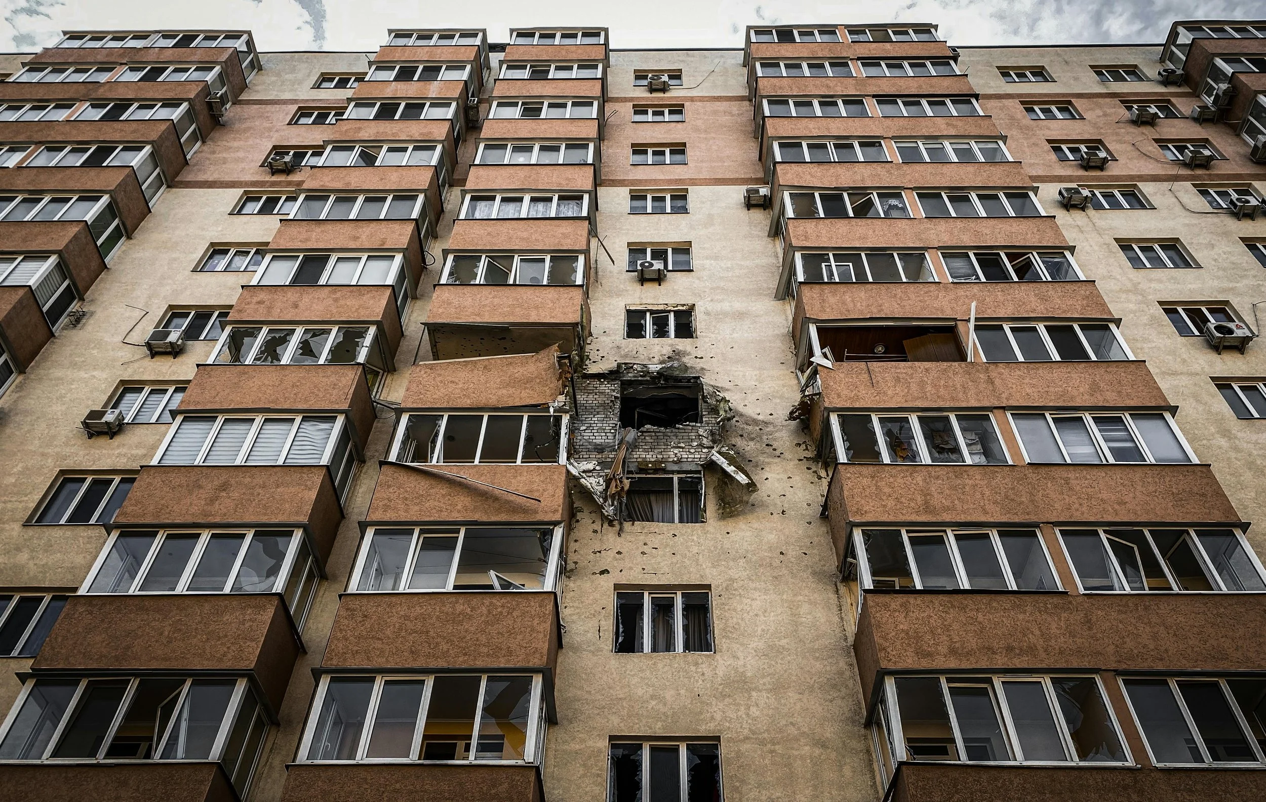 A residential building shows sever damage from an apparent explosion, with debris and shattered windows around the impact zone. Reflecting the perspective of Matt Arlyn on navigating disaster housing programs.