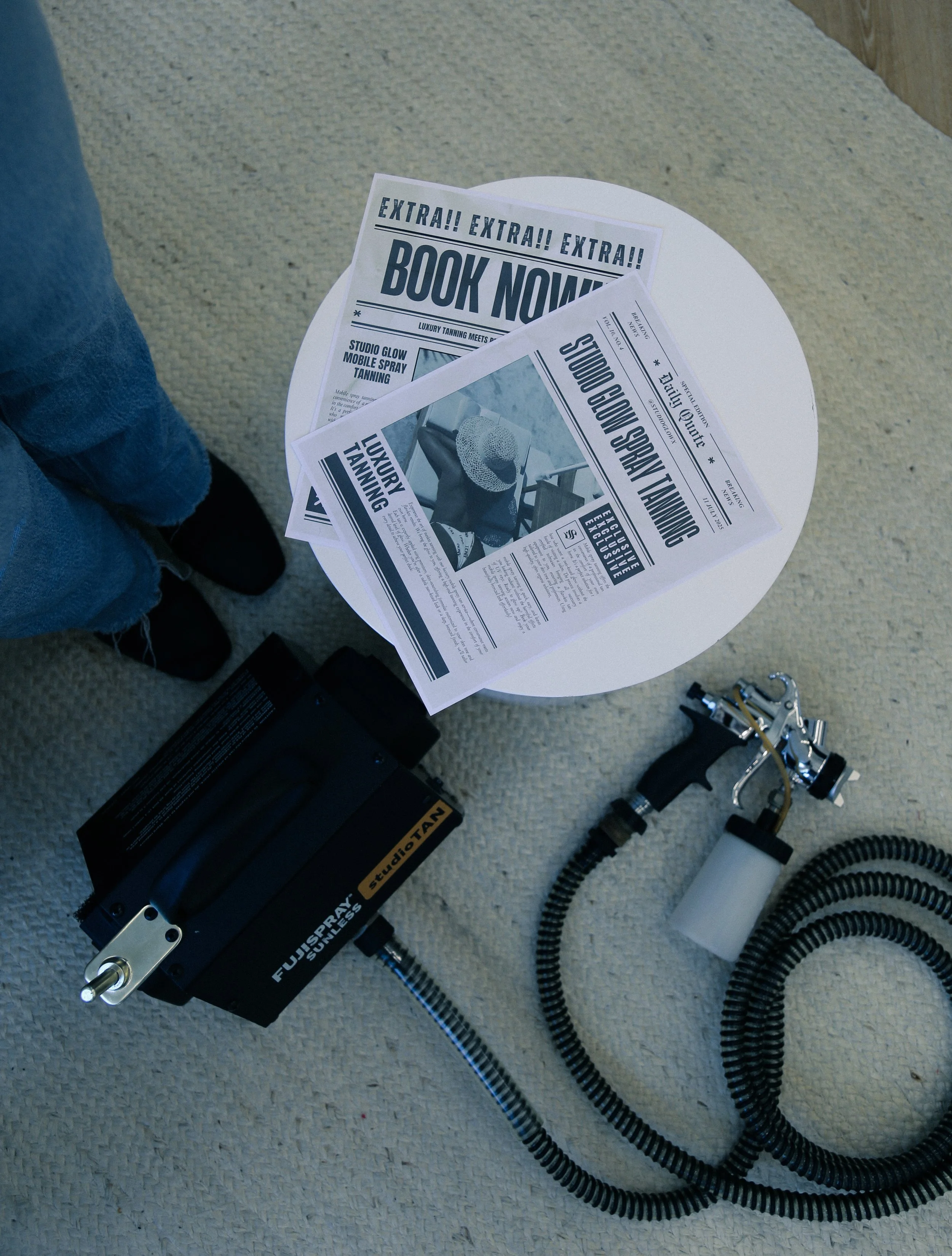 Photograph of a tanning equipment setup with a spray gun, hoses, and posters for tanning services on a small round table, with a person's legs visible nearby.