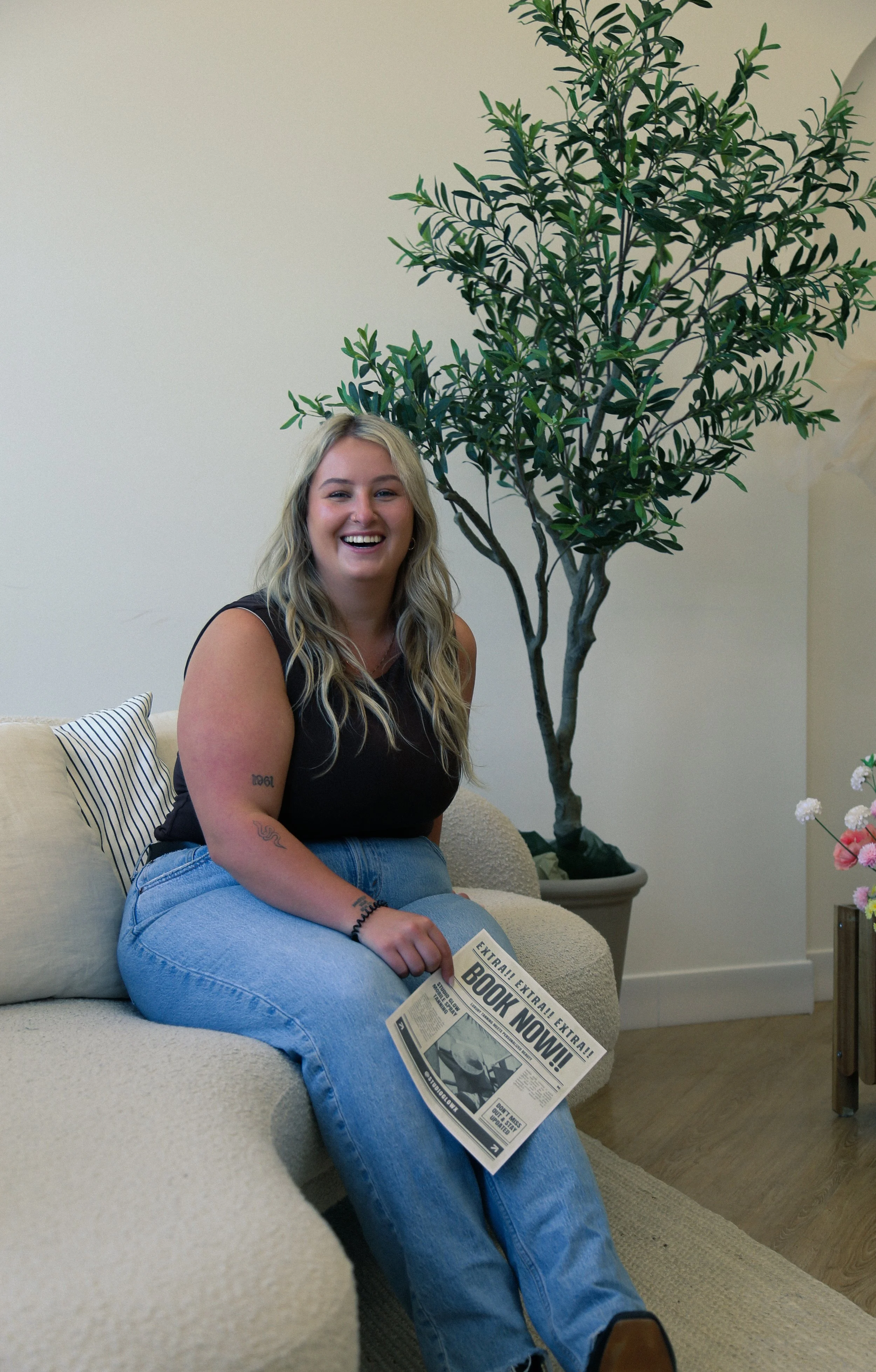 Founder of Studio Glow Tan holding a “Book Now” newspaper in a lifestyle brand photo.
