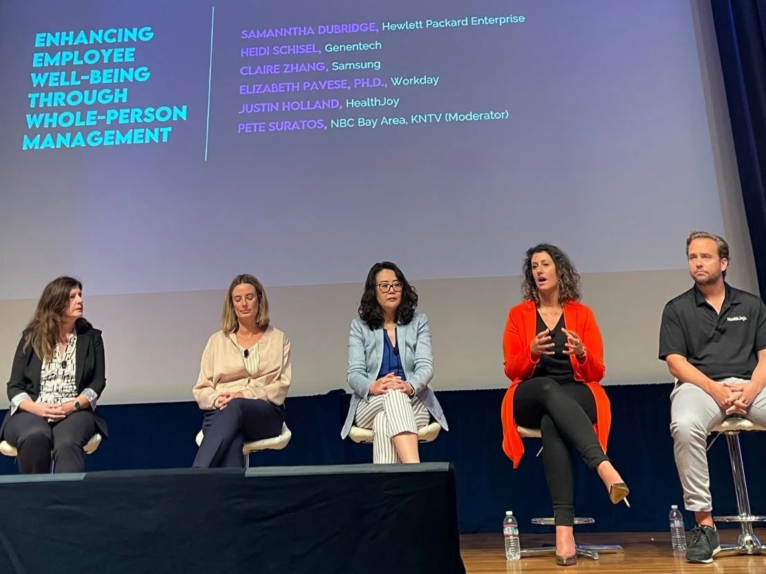 Panel discussion at a conference on enhancing employee well-being through whole-person management. Five panelists, four women and one man, are seated on stage with a large screen behind them displaying the names and affiliations of the speakers.