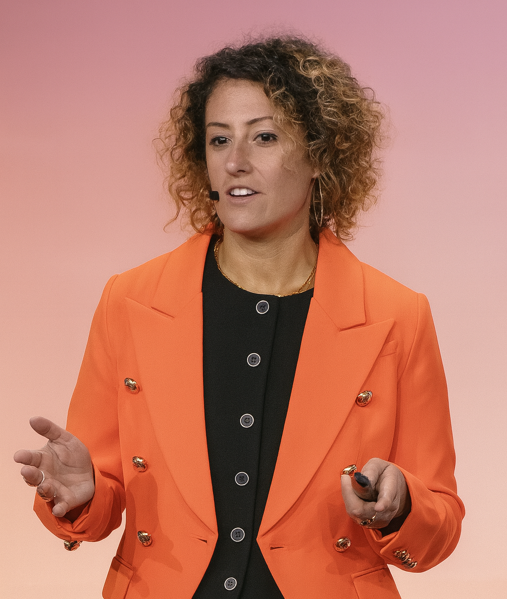 A woman with curly hair wearing an orange blazer and black top, speaking on stage with a pink background.