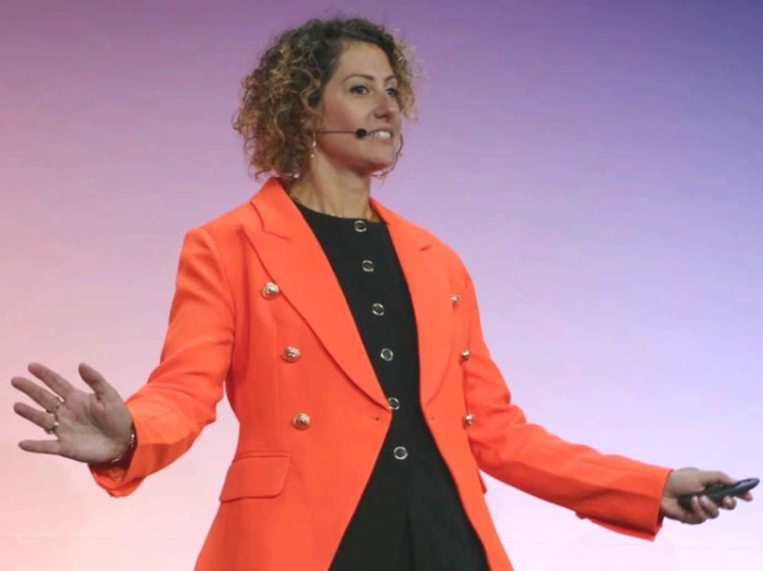 A woman with curly hair giving a presentation on stage, wearing an orange blazer over a black top, holding a remote control in her right hand, with a purple gradient background.