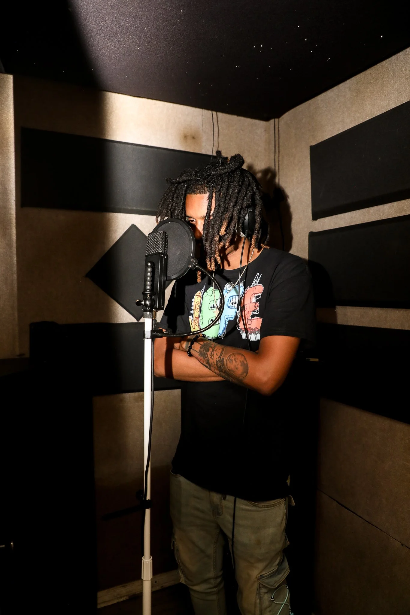 A man with dreadlocks, wearing a black t-shirt with colorful graphics, standing in a recording studio with soundproofing panels, singing or speaking into a microphone.