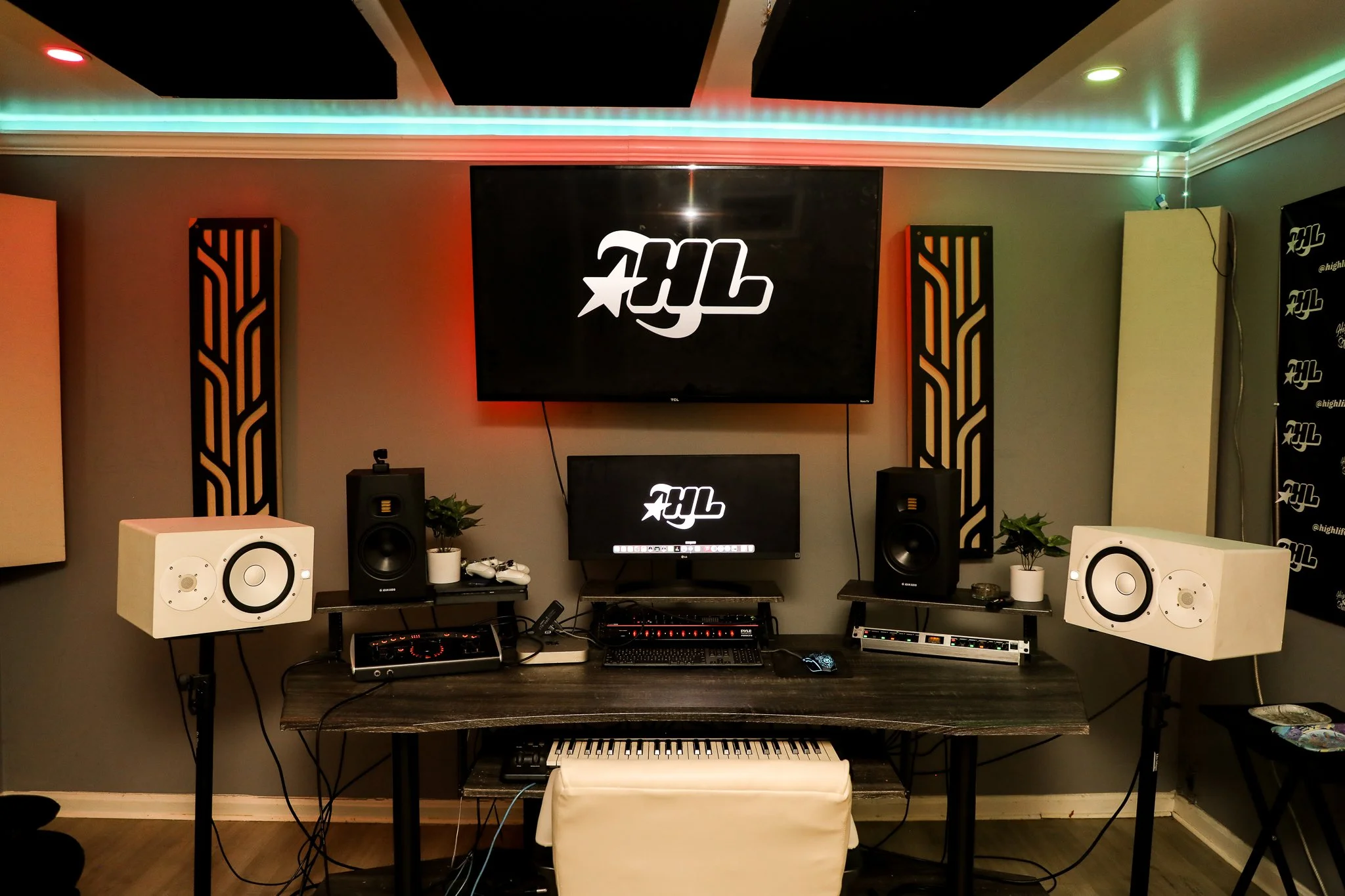 Recording studio with two large white studio monitors, two black studio monitors, a keyboard, a computer, and two TVs displaying a logo, with acoustic panels and colorful LED lighting.