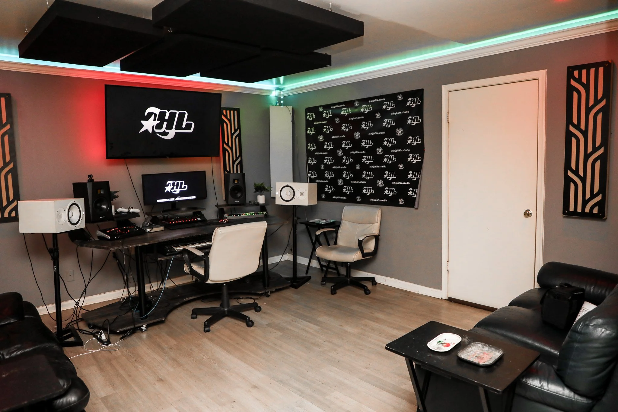 A music studio with wall-mounted speakers, computers, a mixing console, a keyboard, a chair, and a black sofa with a small table.