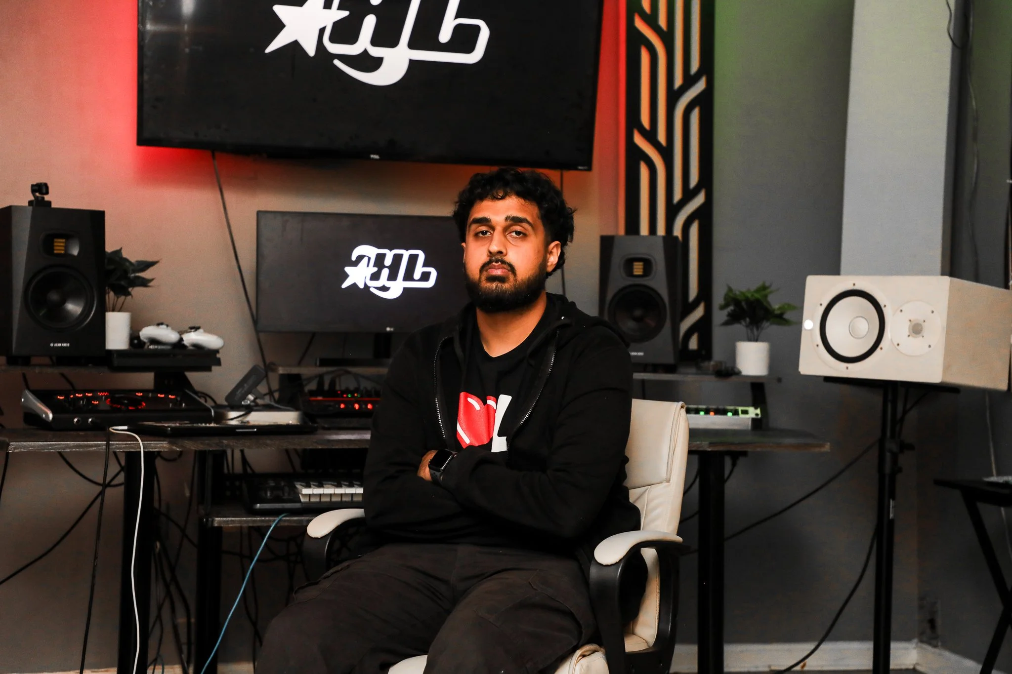 A man with dark hair and beard, wearing a black hoodie with a heart graphic, sits with arms crossed in a music studio surrounded by speakers, monitors, and electronic equipment; large screens behind him display the logo 'FIlX'.