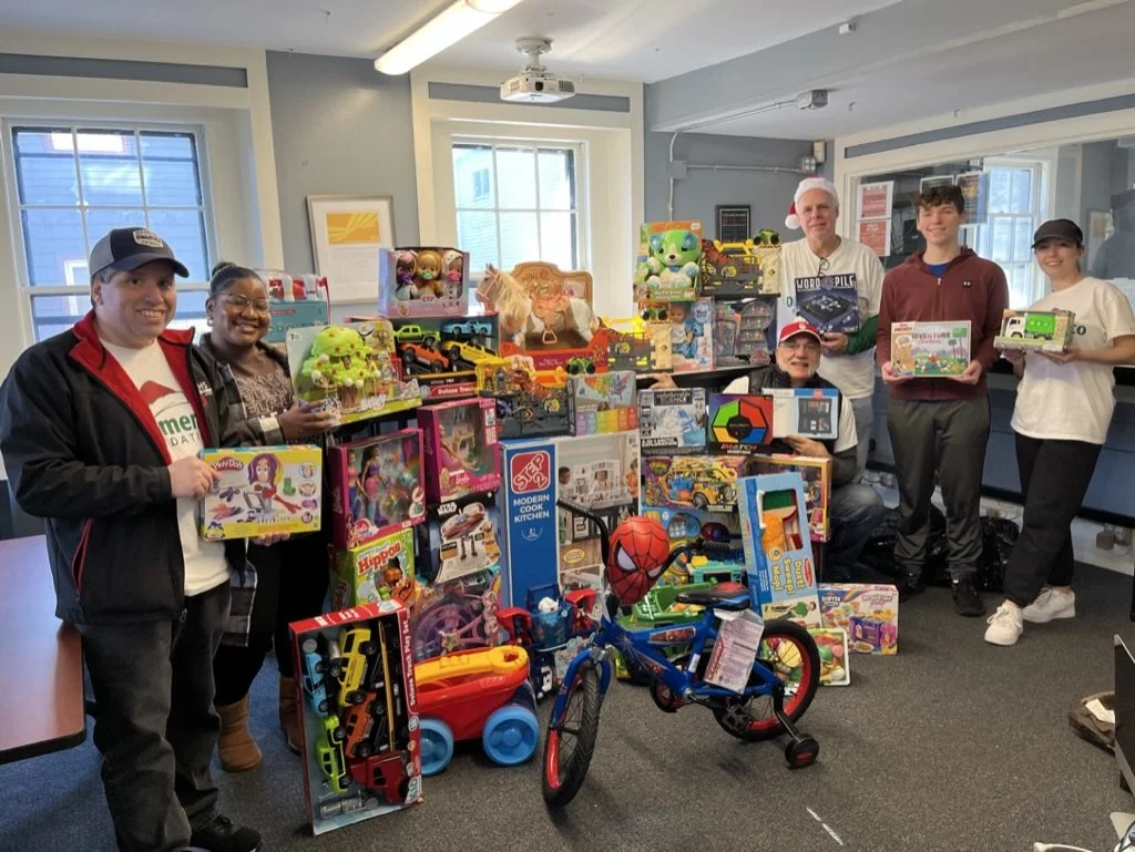Group of people smiling and posing with a large collection of toys, including bikes, dolls, cars, and stuffed animals, in a bright room with windows.