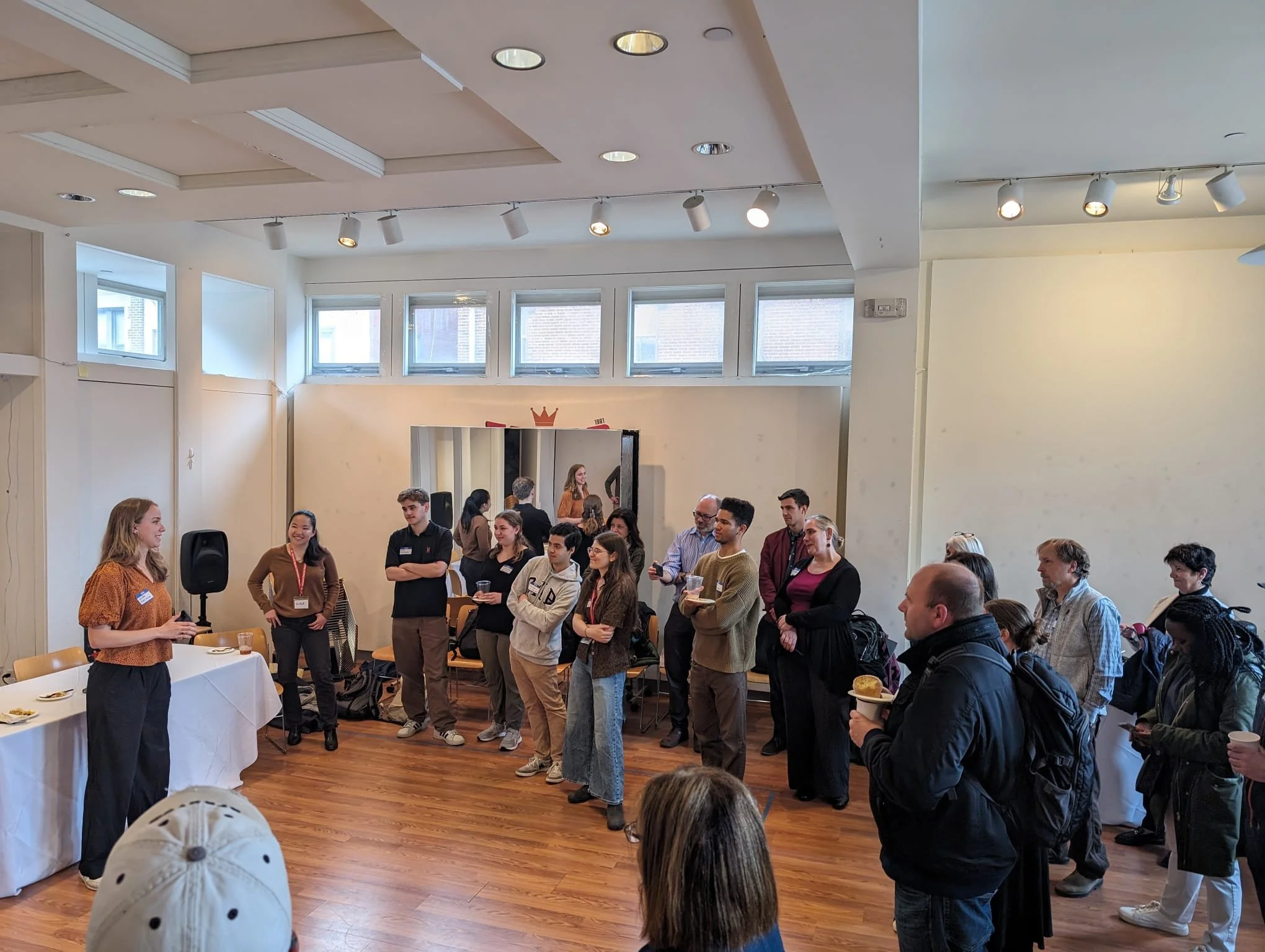A woman giving a presentation to a group of people in a well-lit room with wooden floors. The audience is standing and listening attentively, some holding drinks, with a table on the left side of the image.