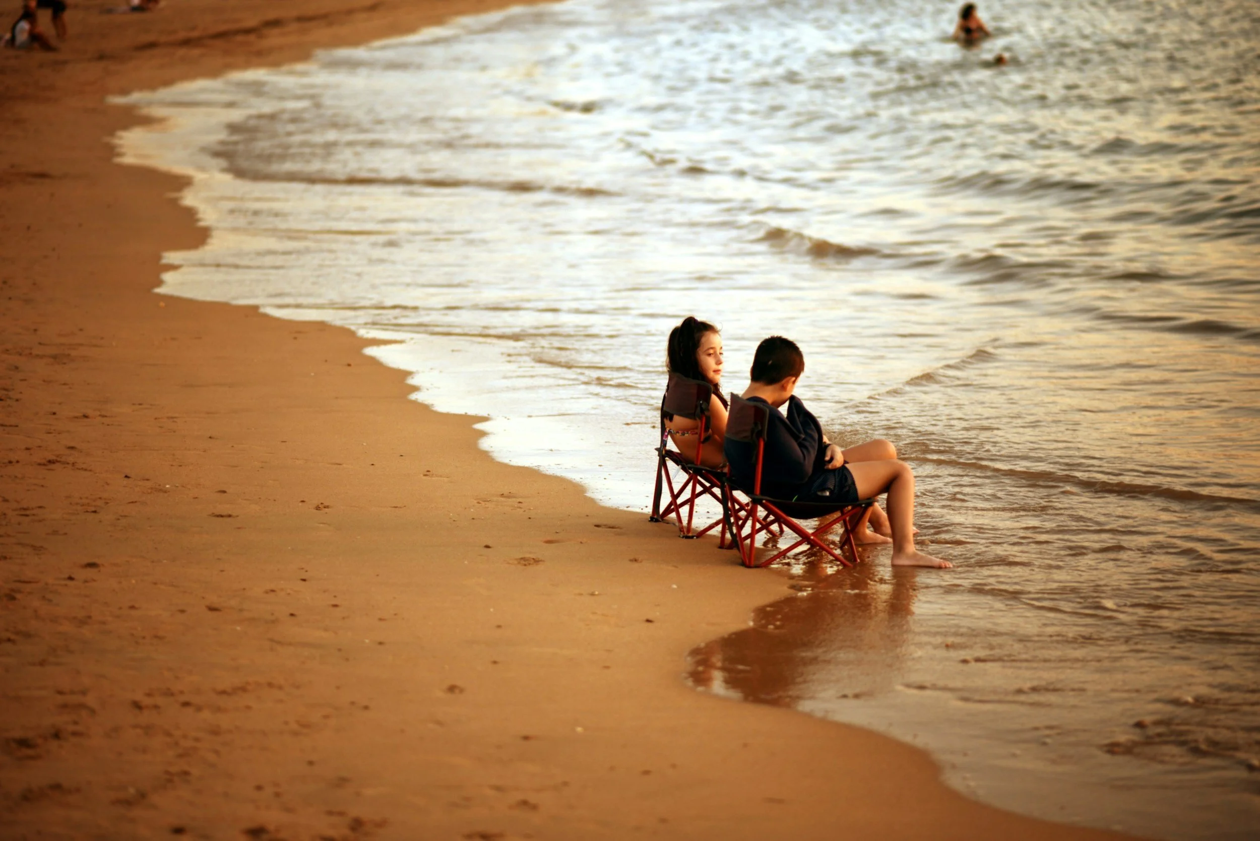 Two children sitting in small portable chairs on the beach, with their feet in the water, during sunset.
