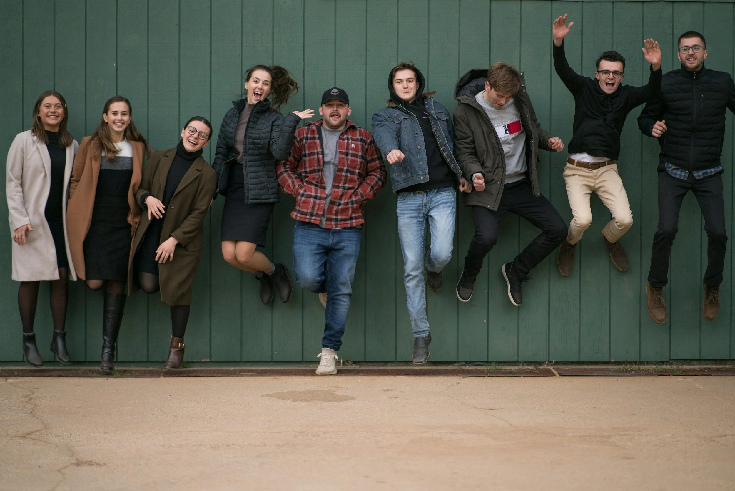 Group of ten young adults standing and jumping in front of a green wall, smiling and enjoying themselves.