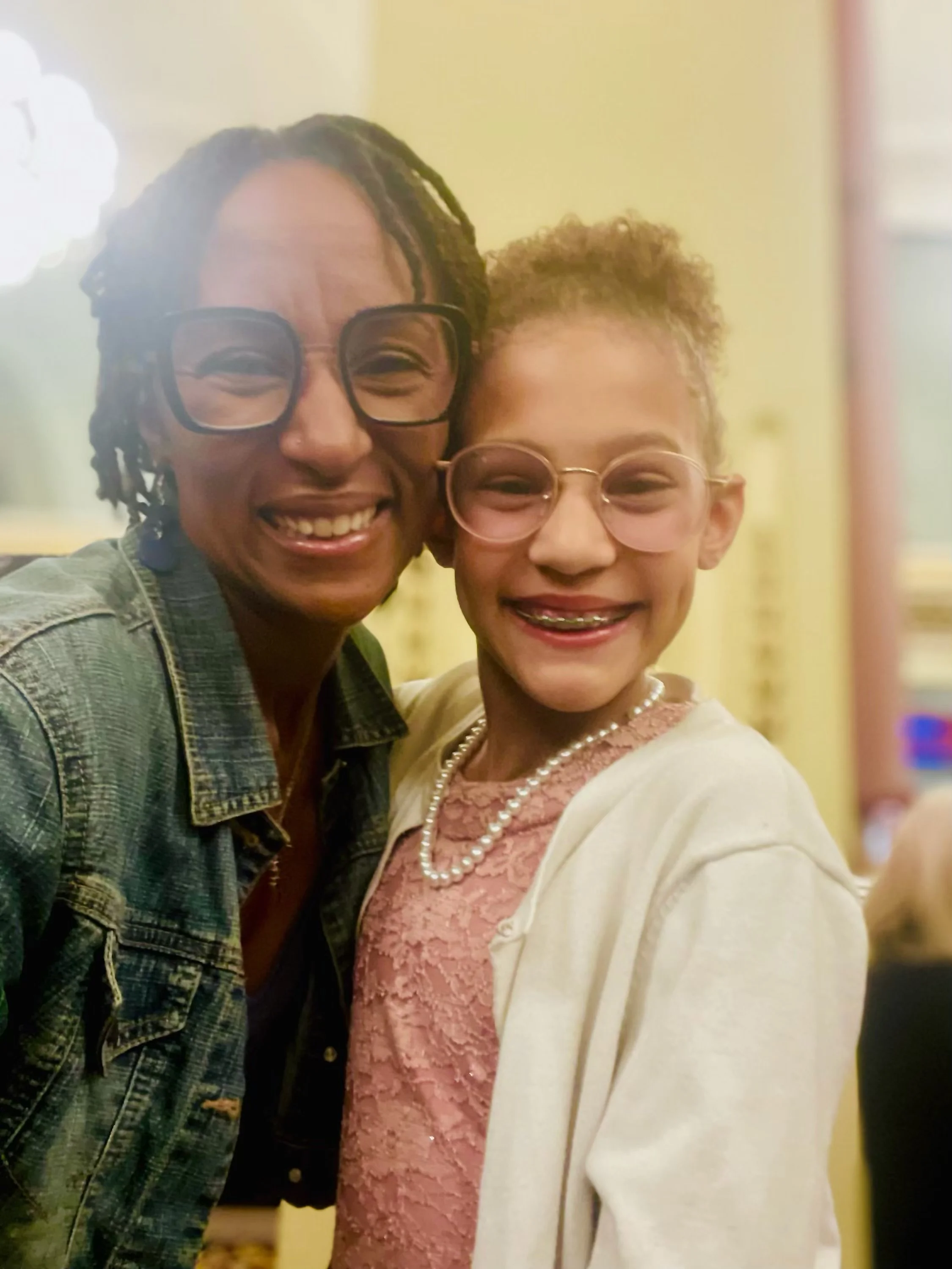 A smiling woman with glasses and dreadlocks taking a selfie with a smiling young girl with glasses and braces, both dressed nicely, in an indoor setting.