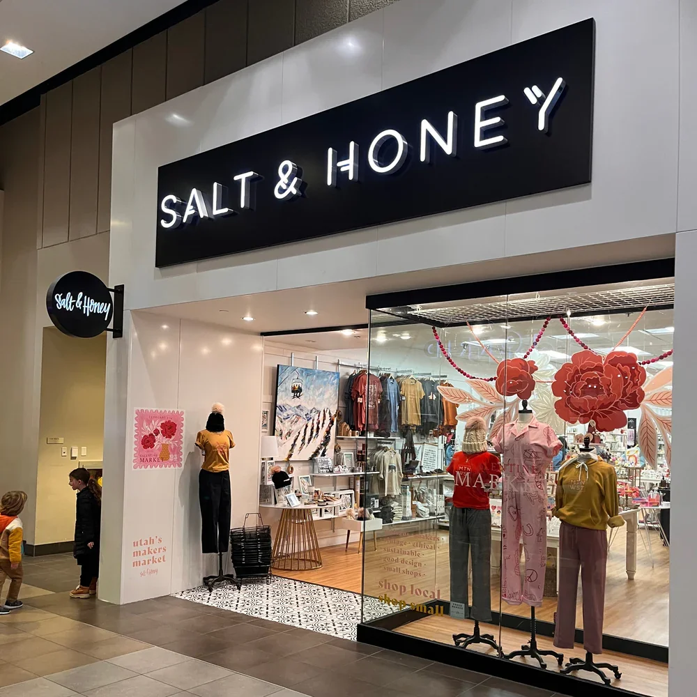 Storefront with sign 'Salt & Honey' and mannequins displaying clothing inside. Two children standing outside looking in, with other shoppers inside visible through large glass display window.
