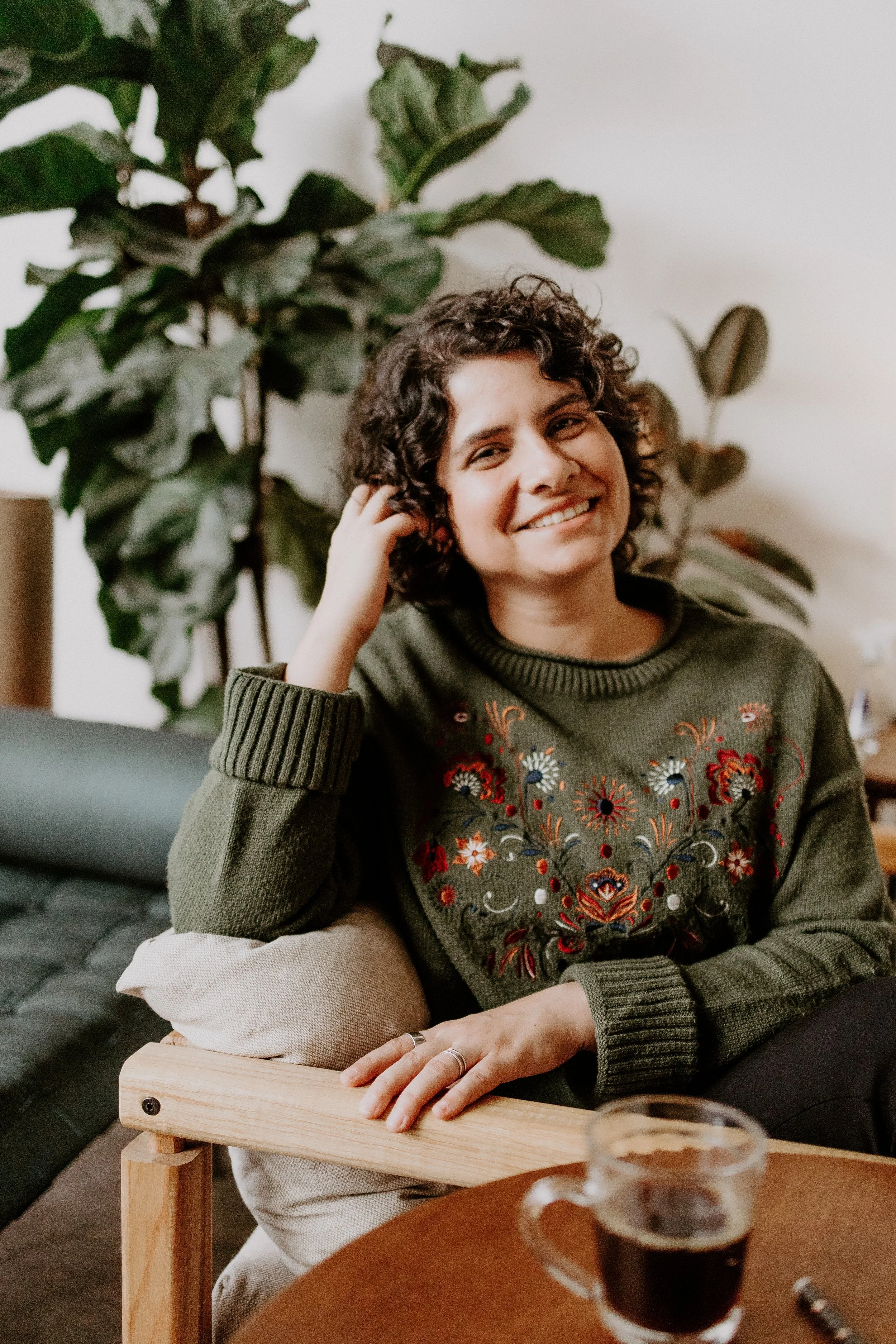 Mulher sorridente com cabelo cacheado sentada em uma sala, usando suéter verde com estampa floral.